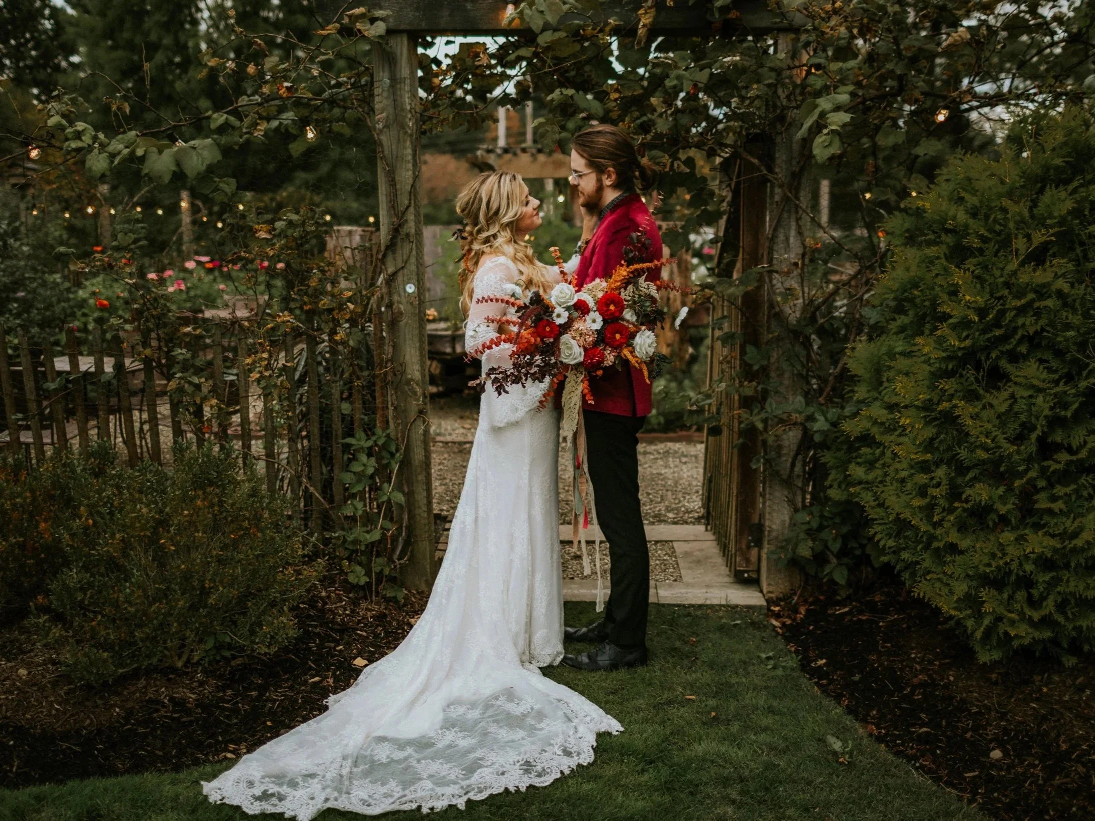 A couple stands together outdoors under a tree with string lights, during a wedding ceremony. The woman is in a white lace wedding dress holding a colorful bouquet, and the man is in a red blazer with black pants. They are looking into each other's eyes.