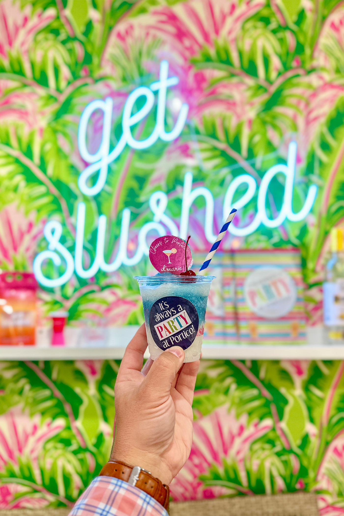 A hand holding a colorful frozen drink at a party-themed location with a bright neon sign that says "get lit" and lush tropical floral wallpaper in the background.
