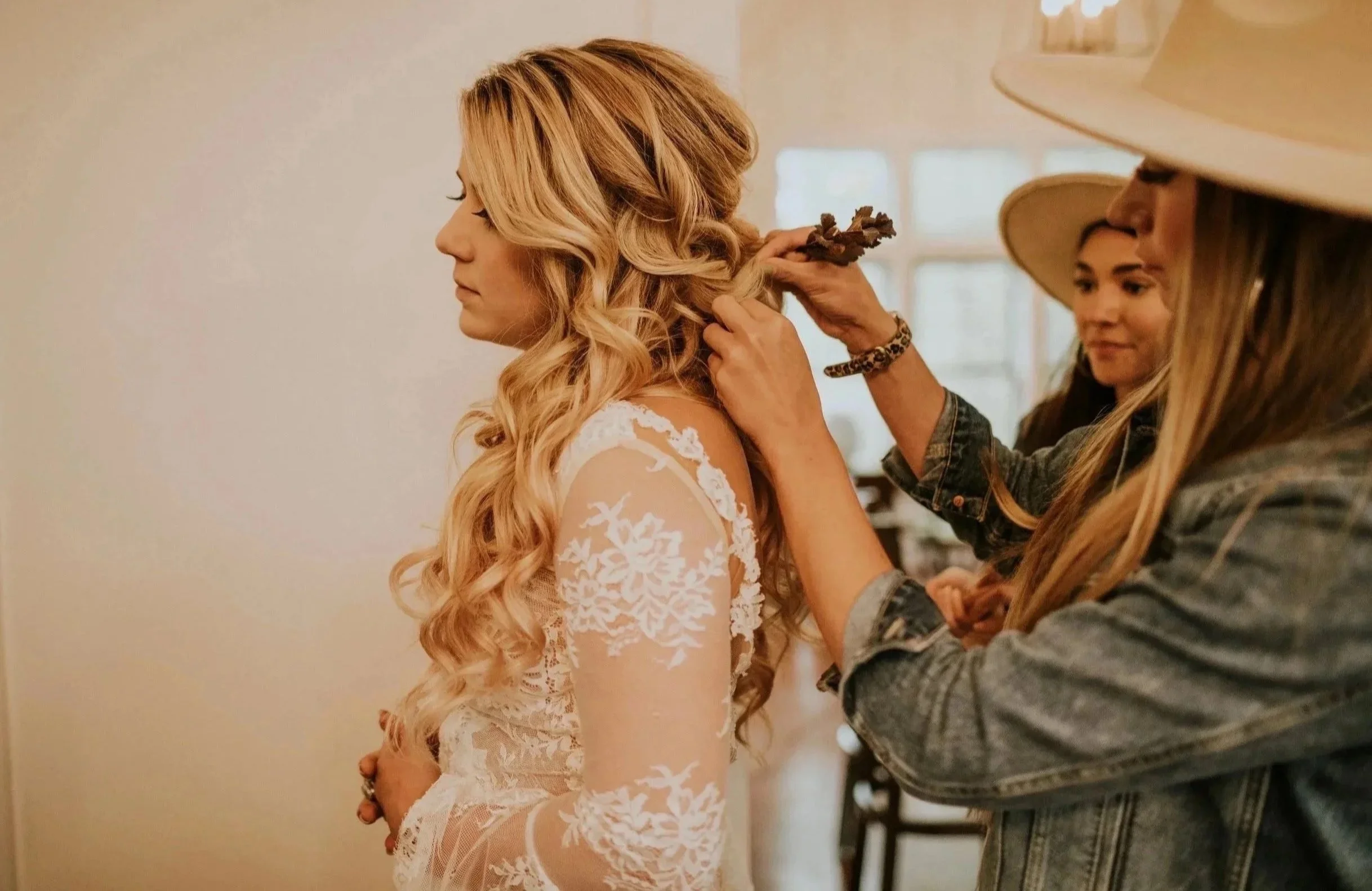A woman with long, curly blonde hair is having her hair styled by another woman who is wearing a cowboy hat. The woman getting her hair styled is wearing a white lace dress.