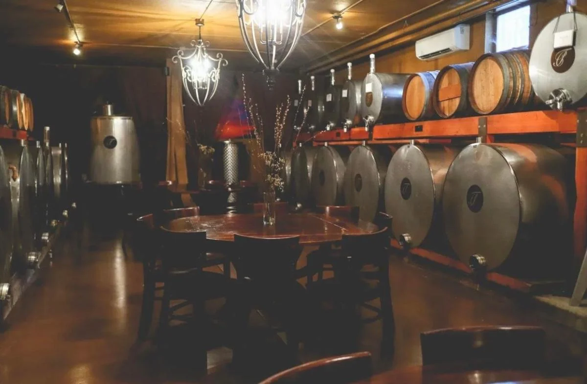 Interior of a winery or brewery with wine barrels and equipment, a wooden table with chairs, chandelier, wooden walls, and an air conditioning unit.