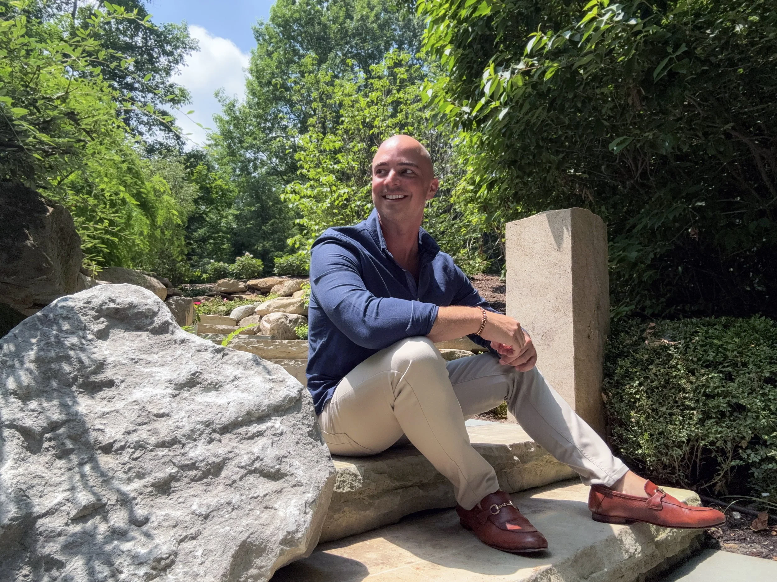 A man with a shaved head wearing a blue shirt and white pants sitting outdoors on rocks surrounded by greenery, with a smile on his face and#39; looking to the side. He is near a stone block and appears relaxed.
