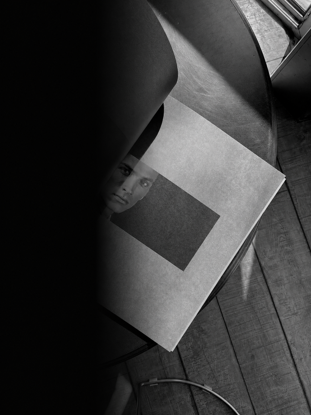 Black and white photo of an open magazine on a wooden table, with a woman's face on the page, partially obscured, and a window in the background.