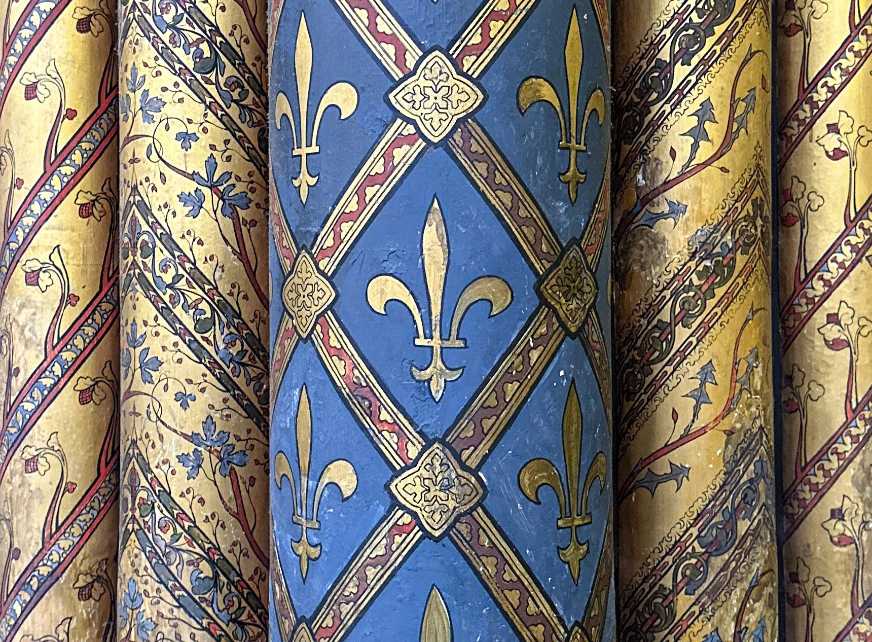 Close-up of a decorative, colorful, patterned surface with gold, red, black, and blue designs featuring fleur-de-lis symbols, floral motifs, and intricate geometric lines.