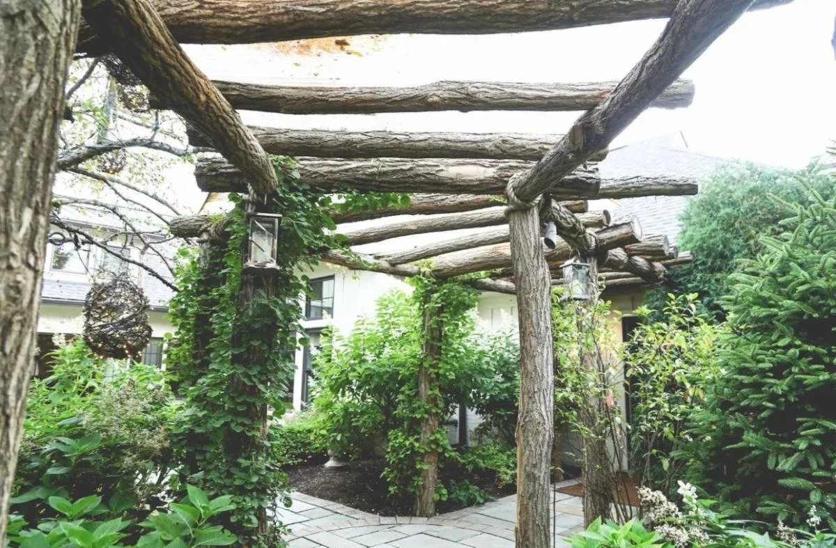 Wooden pergola with rustic logs in a lush green garden, hanging lanterns, and a stone patio.