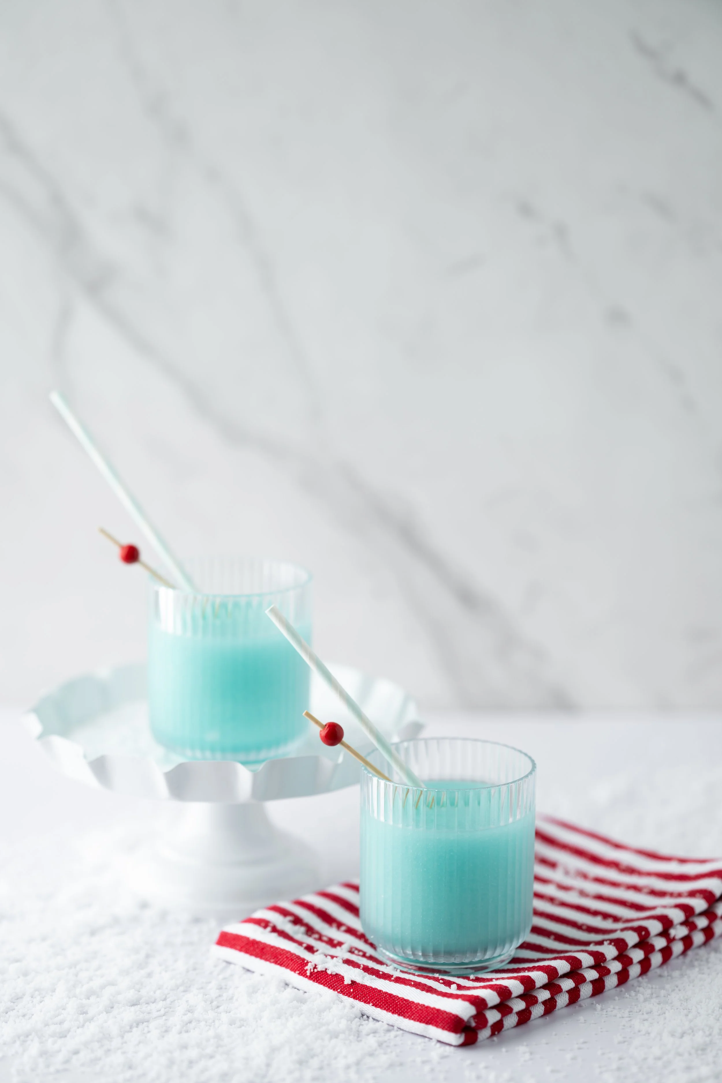 Two clear glasses filled with blue-colored beverages, each with a straw featuring a red cherry on top. The glasses are placed on a red and white striped cloth on a white textured surface, with a white paper tray underneath. There is a white background with a subtle marble pattern.