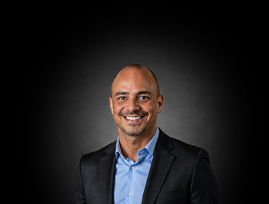 Portrait of a smiling man with a shaved head, wearing a black suit jacket and a light blue collared shirt, against a dark background.