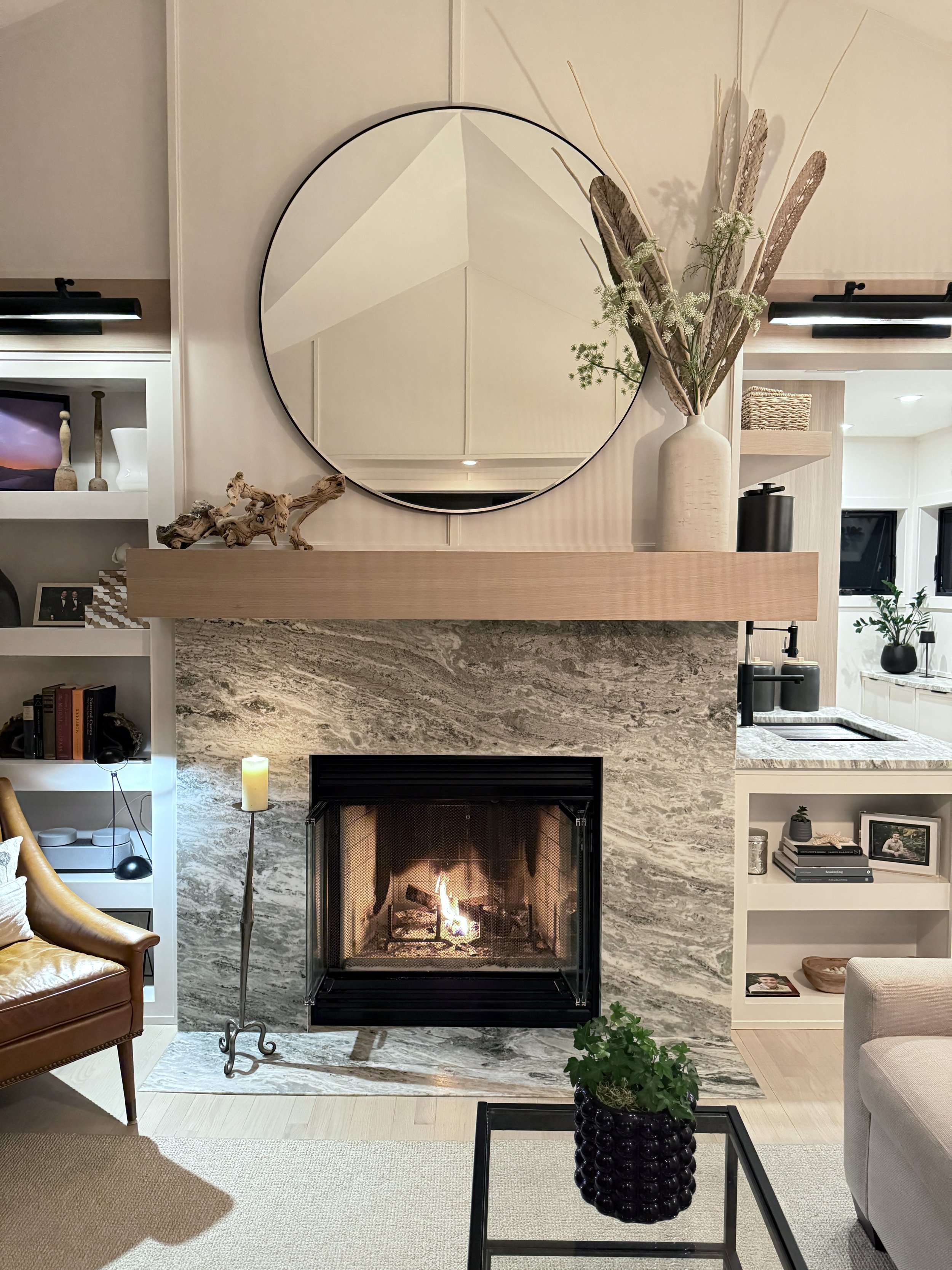 Living room with a fireplace, a large circular mirror above the mantel, a tall white vase with dried plants, a potted plant on a black table, a beige armchair, a brown armchair, and shelves with decorative objects and books.