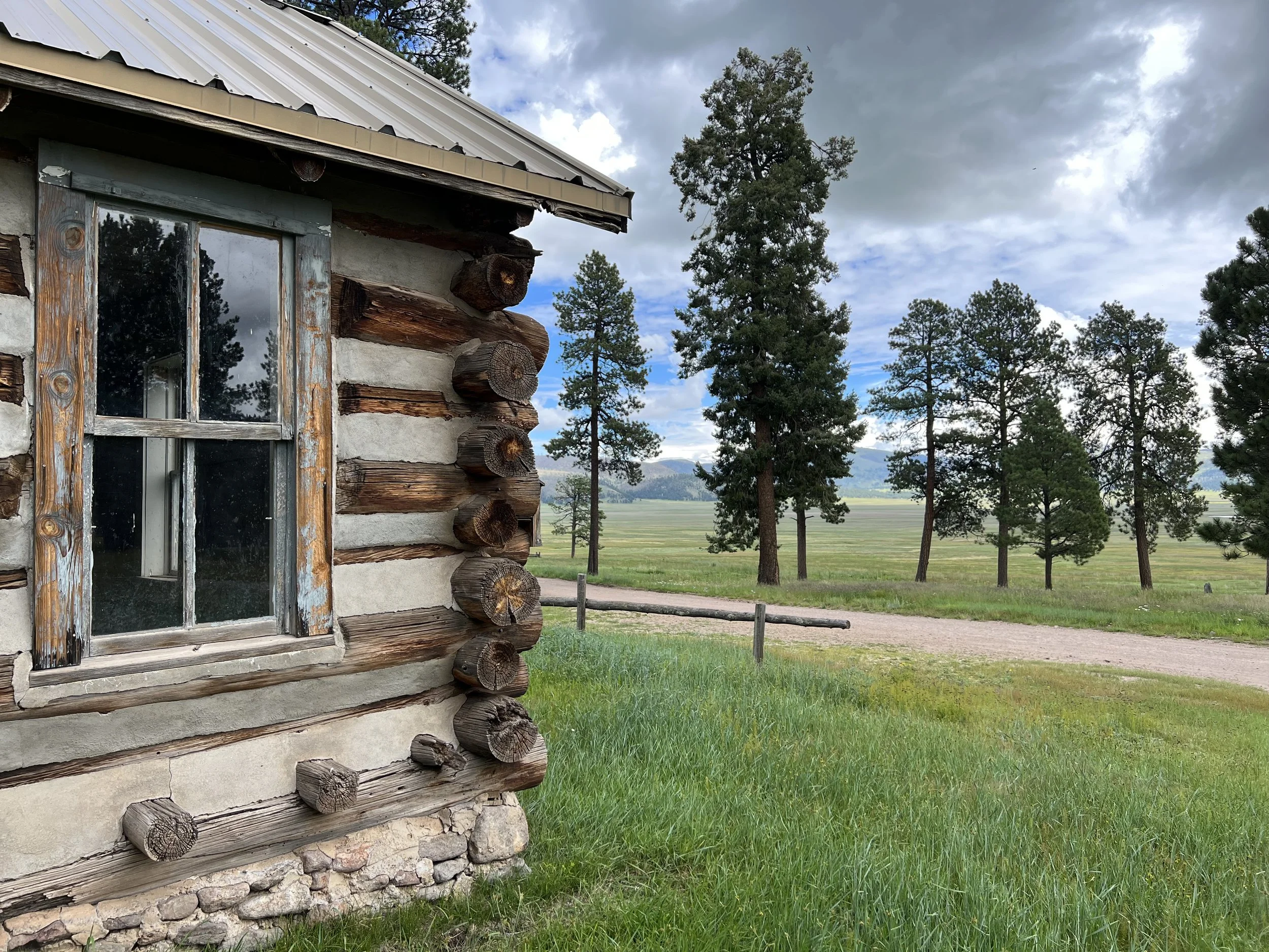 Side of a log cabin with wooden logs and a window, set in a landscape with tall trees and a cloudy sky.