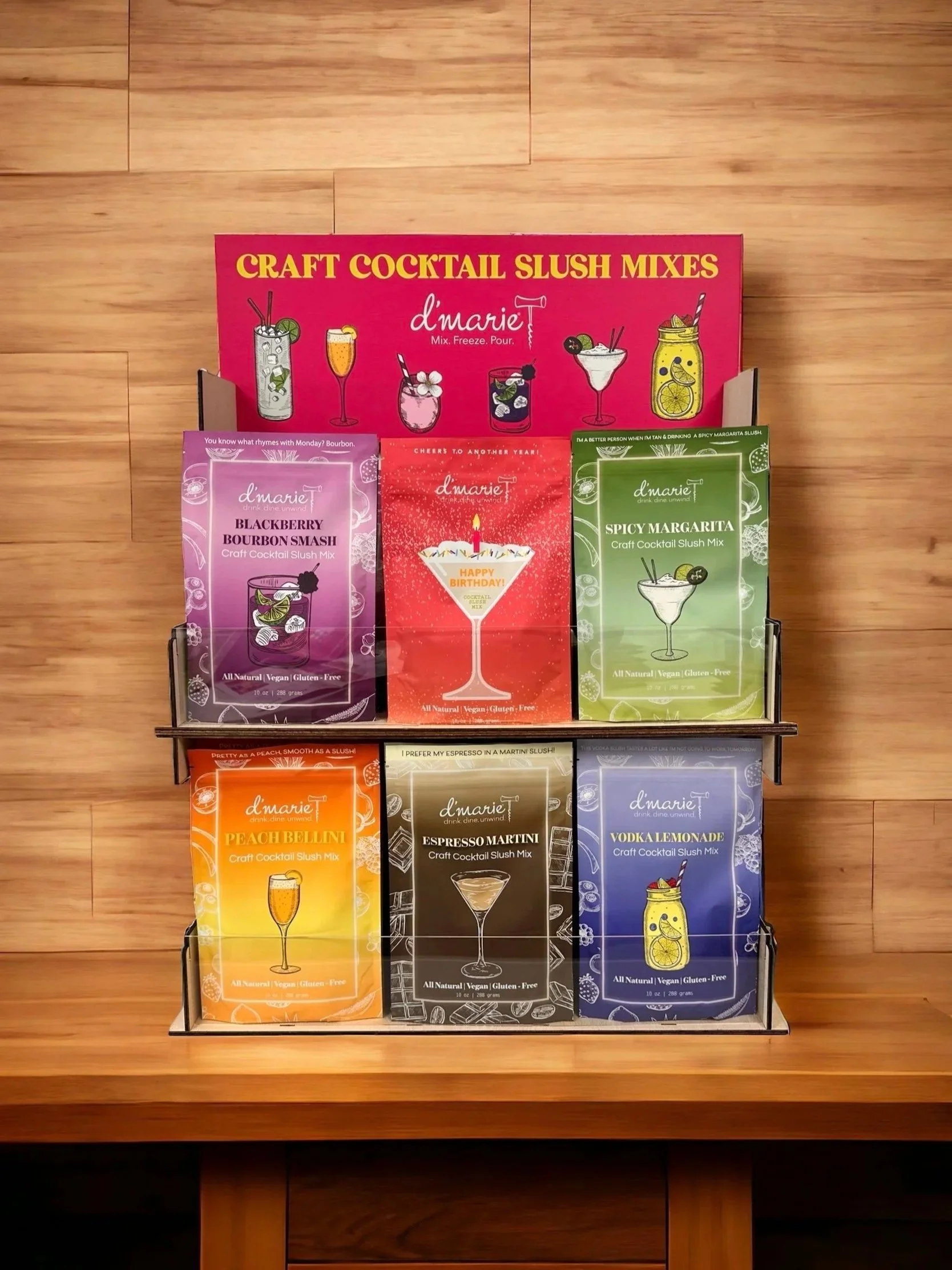 Display stand with colorful packages of craft cocktail slush mixes, including Blackberry Bourbon Smash, Happy Birthday, Spicy Margarita, Peach Bellini, Espresso Martini, and Vodka Lemonade, on a wooden surface.