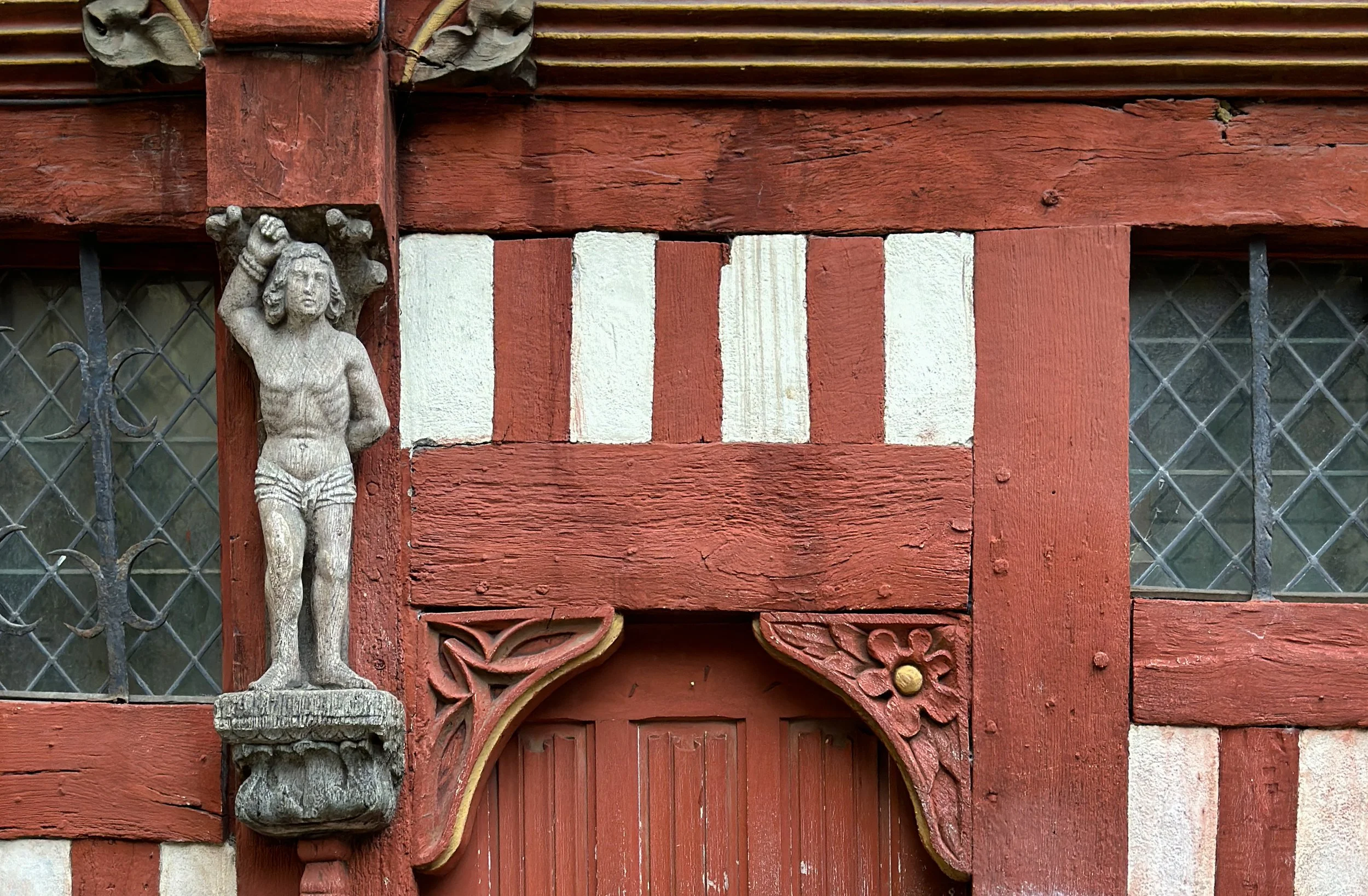 Decorative wooden structure with a small carved female figure and a diamond-patterned metal grid window in the background.