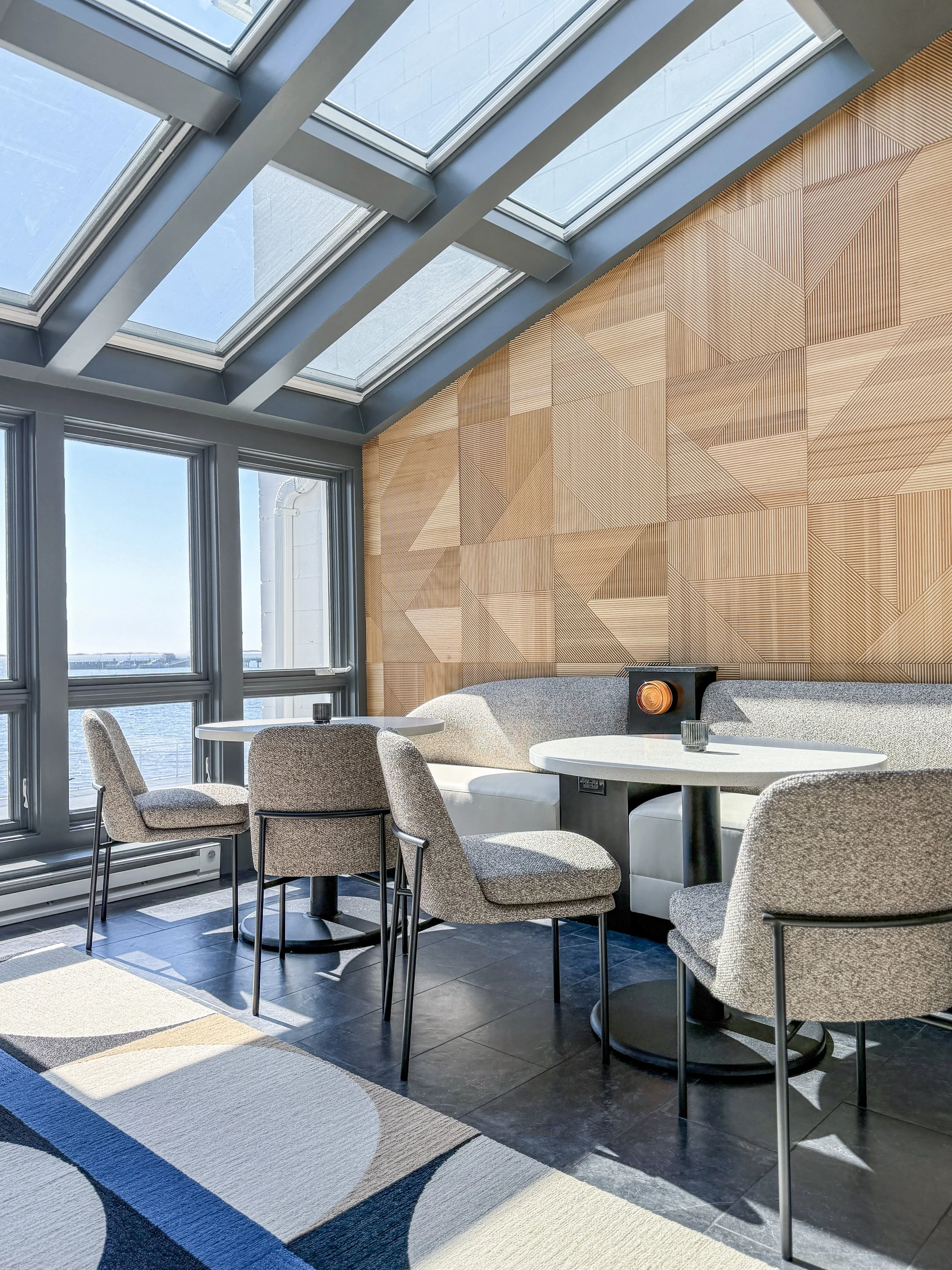 A modern dining area with round tables, upholstered chairs, a wooden accent wall, large windows with a view of water, and a ceiling with skylights.