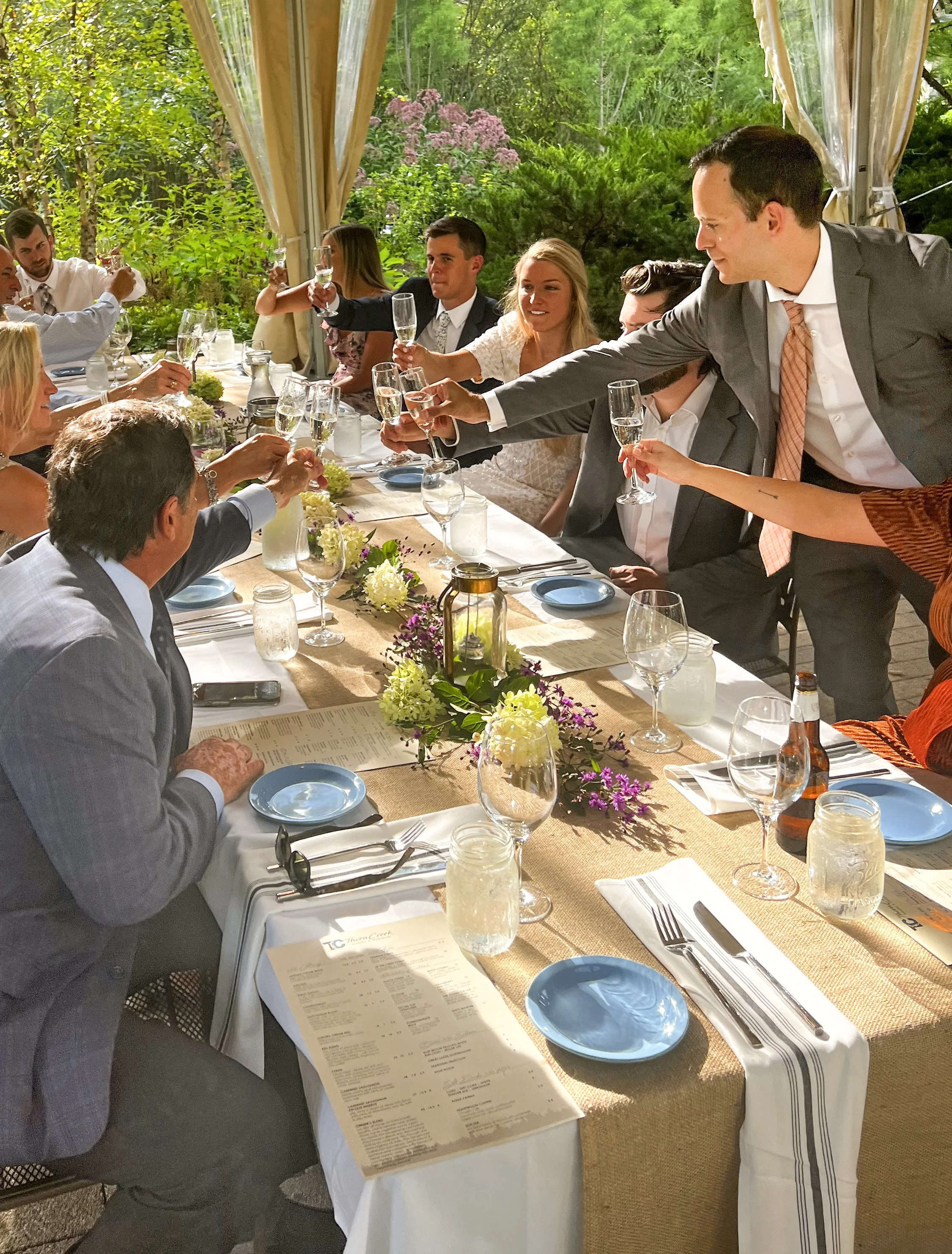 People at a long dining table raising glasses in a toast at an outdoor event with lush green trees in the background, decorated with flowers and table settings.