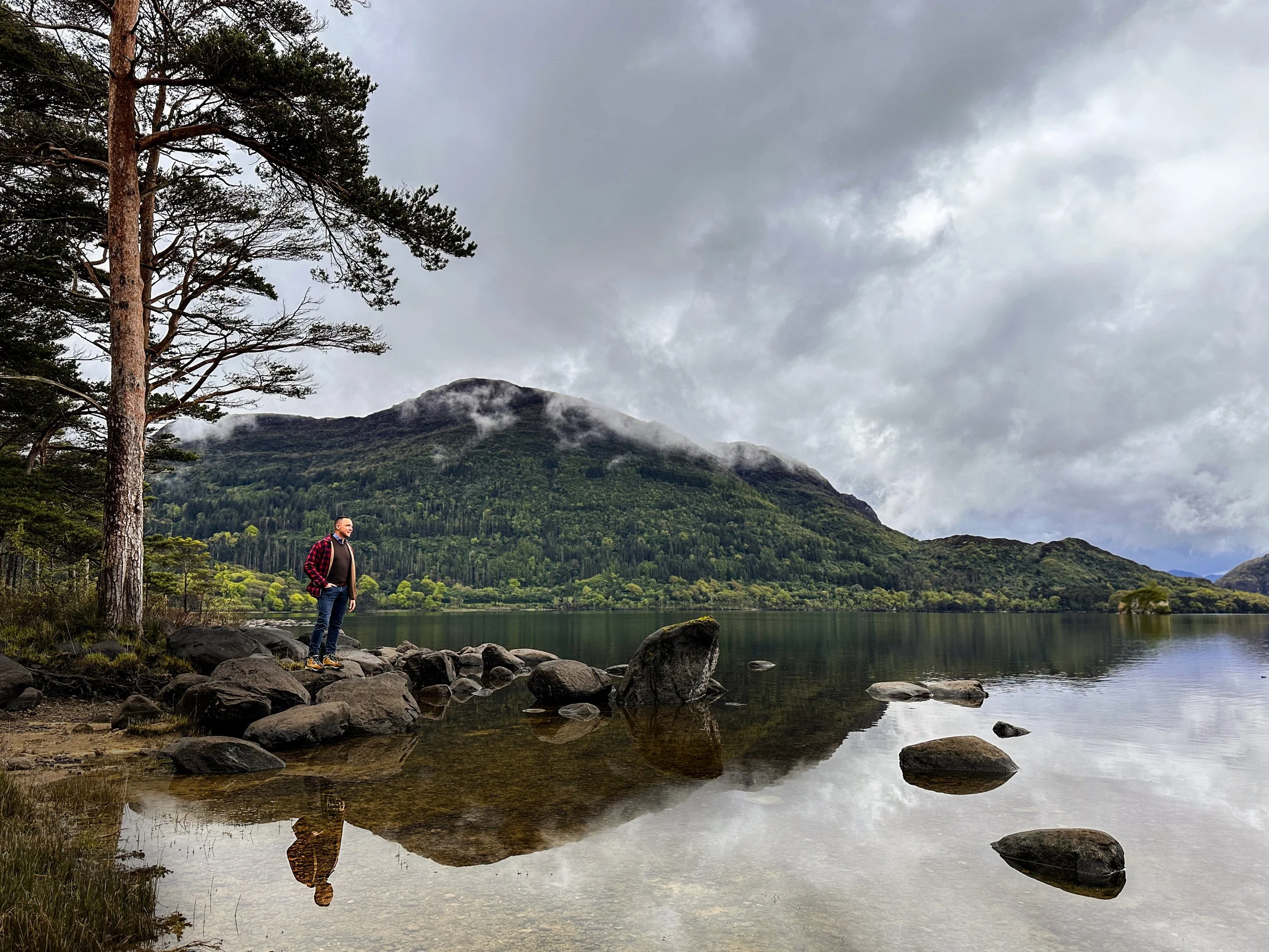 A man in a red checkered jacket standing on rocks near a calm lake, with mountains and cloudy sky in the background.