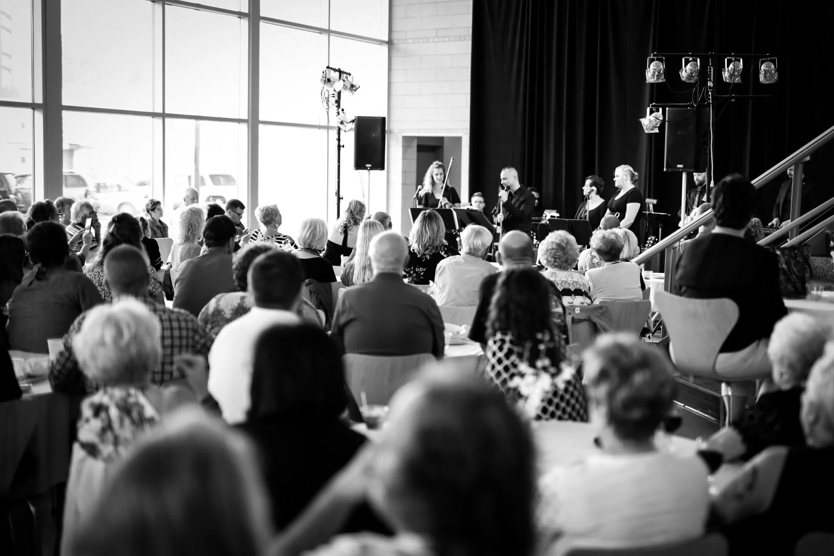 A black and white photo of a musical performance with a singer and violinist on stage, in front of an audience in a large, modern room with floor-to-ceiling windows and stage lighting.