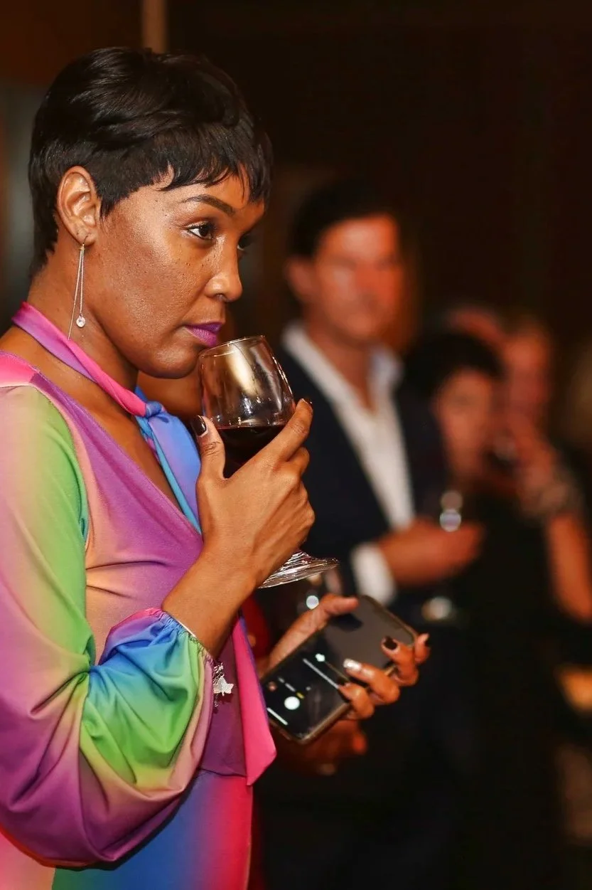 Woman with short dark hair and earrings holding a glass of red wine while looking at her smartphone at a social event.