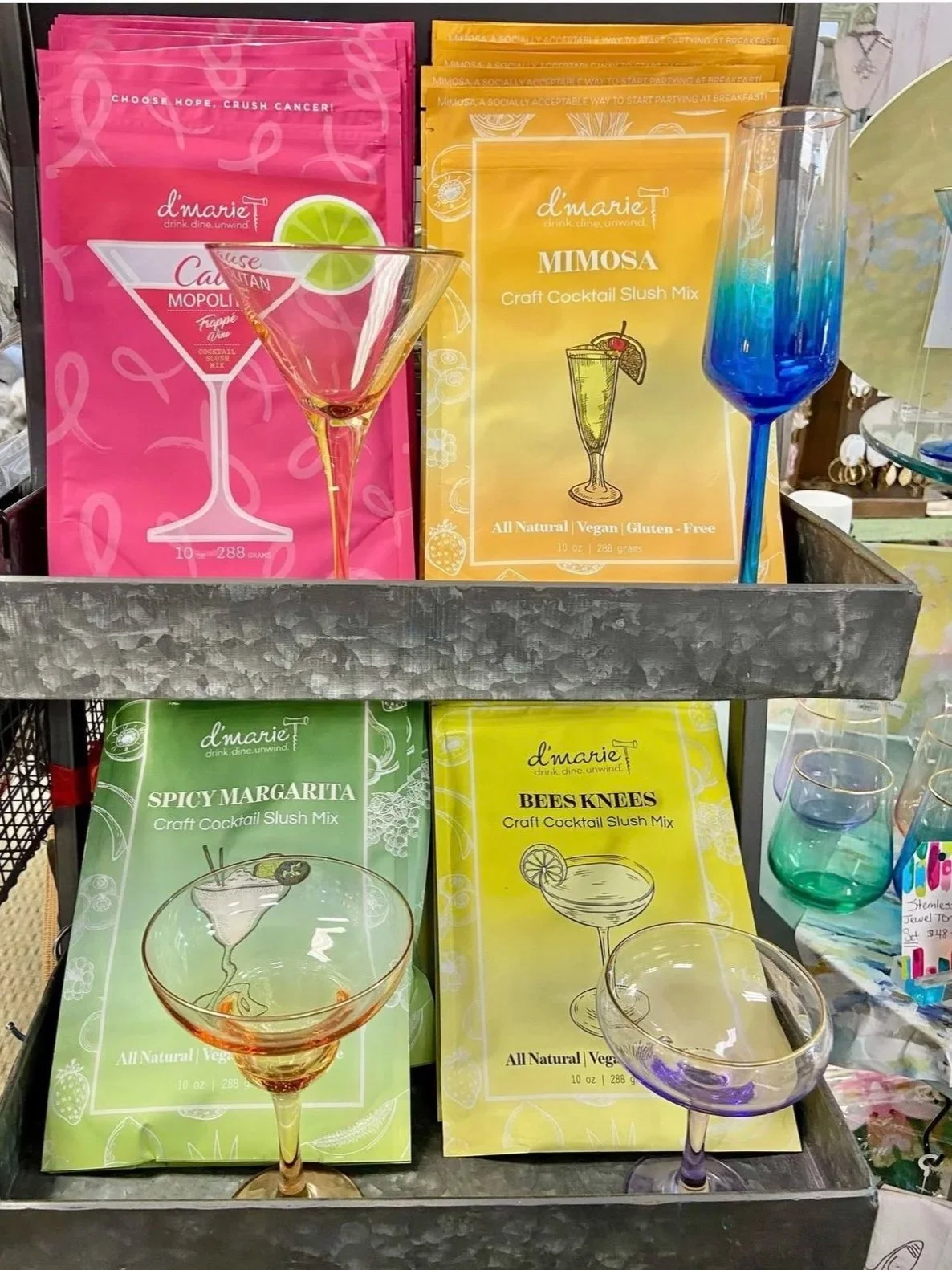 Display of four packets of cocktail slush mixes in colorful packaging and clear plastic drinkware on a metal shelf.
