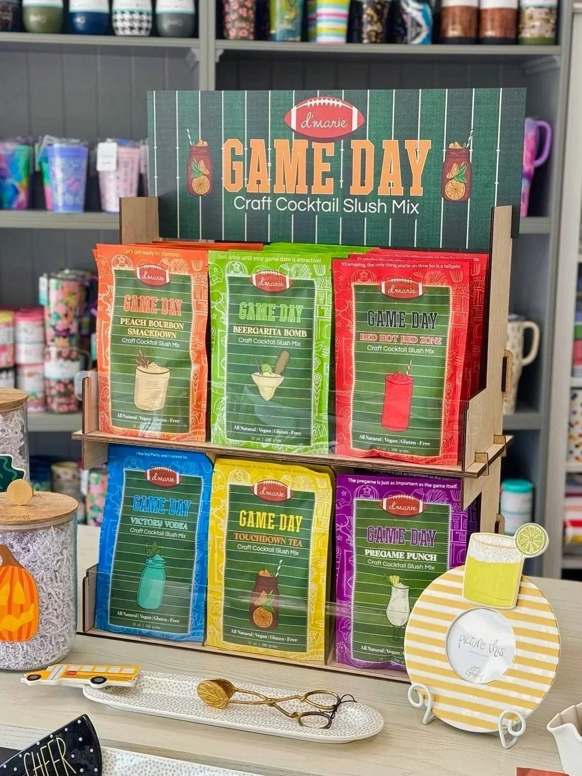 Display of craft cocktail slush mixes called Game Day by d'marie, featuring flavors such as Peach Bourbon Smackdown, Beergarita Bomb, Red Hot Red Zone, Victory Vodka, Touchdown Tequila, and Pregame Punch, arranged on a wooden stand with a background of shelves filled with colorful cups.