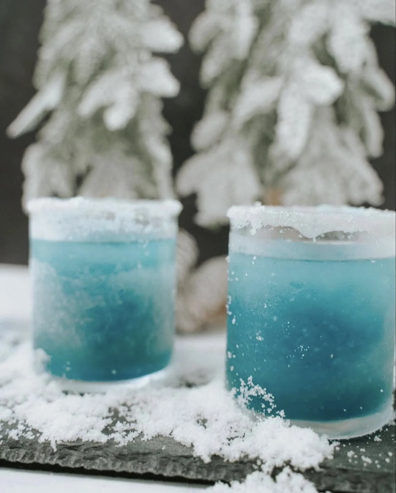 Two blue candles in glass holders placed on a snow-covered surface with snow-covered evergreen trees in the background.