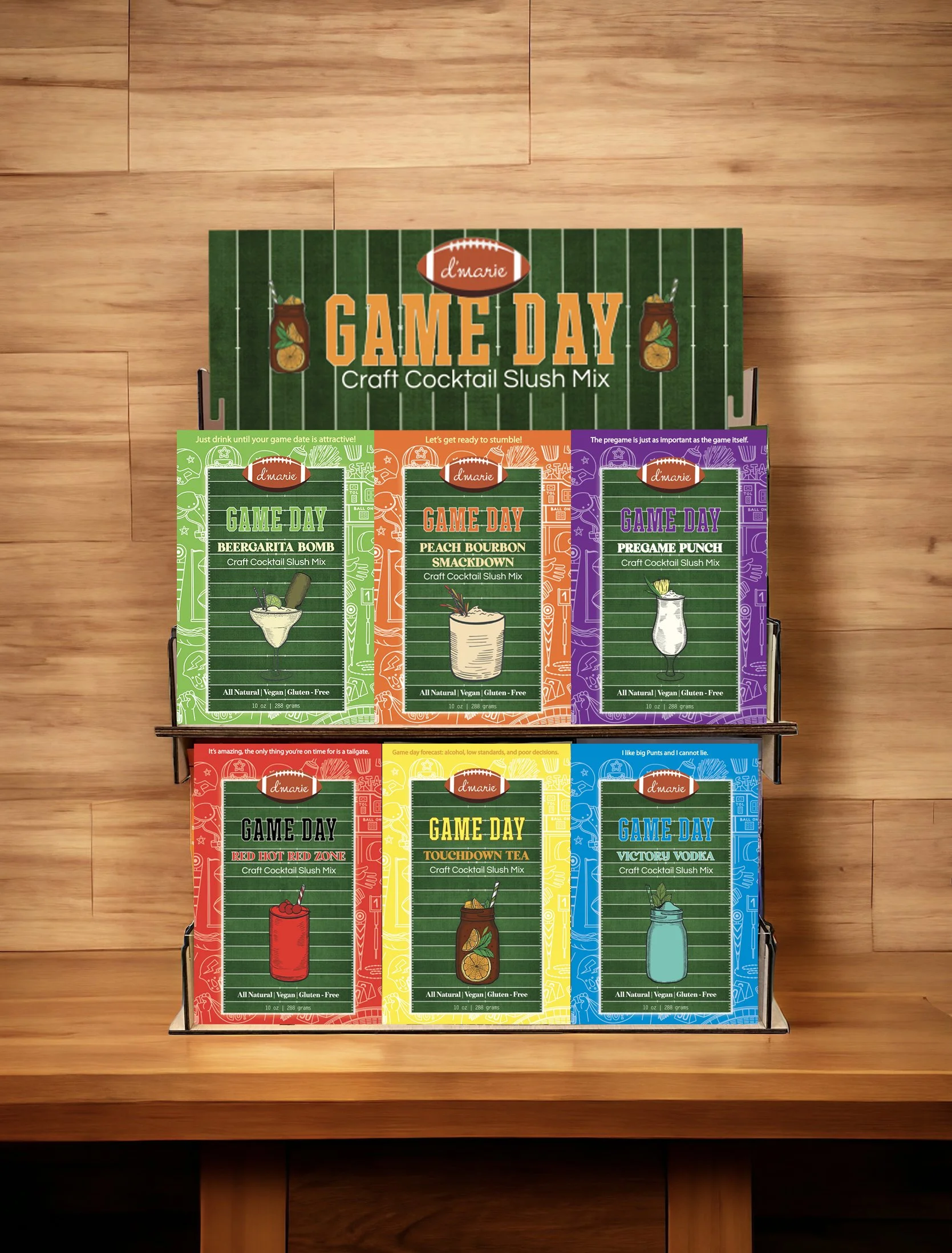 Display of craft cocktail slush mix packets with a green football field background theme, labeled 'Game Day' with different flavors including Beergarita Bomb, Peach Bourbon Smackdown, PreGame Punch, Red Hot Red Zone, Touchdown Tea, and Victory Vodka, arranged in two rows on a wooden surface.