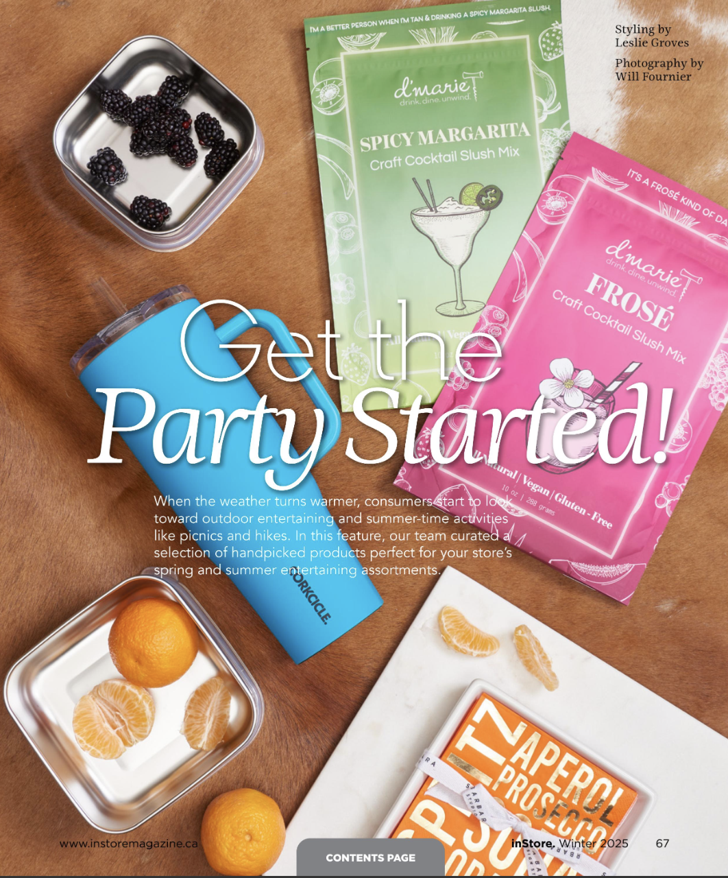 Magazine page featuring summer party items such as blackberries, clementines, drink mix packets labeled 'Spicy Margarita' and 'Frose', a blue plastic wine sippy cup, and white marbled tray with orange slices, on a brown table.