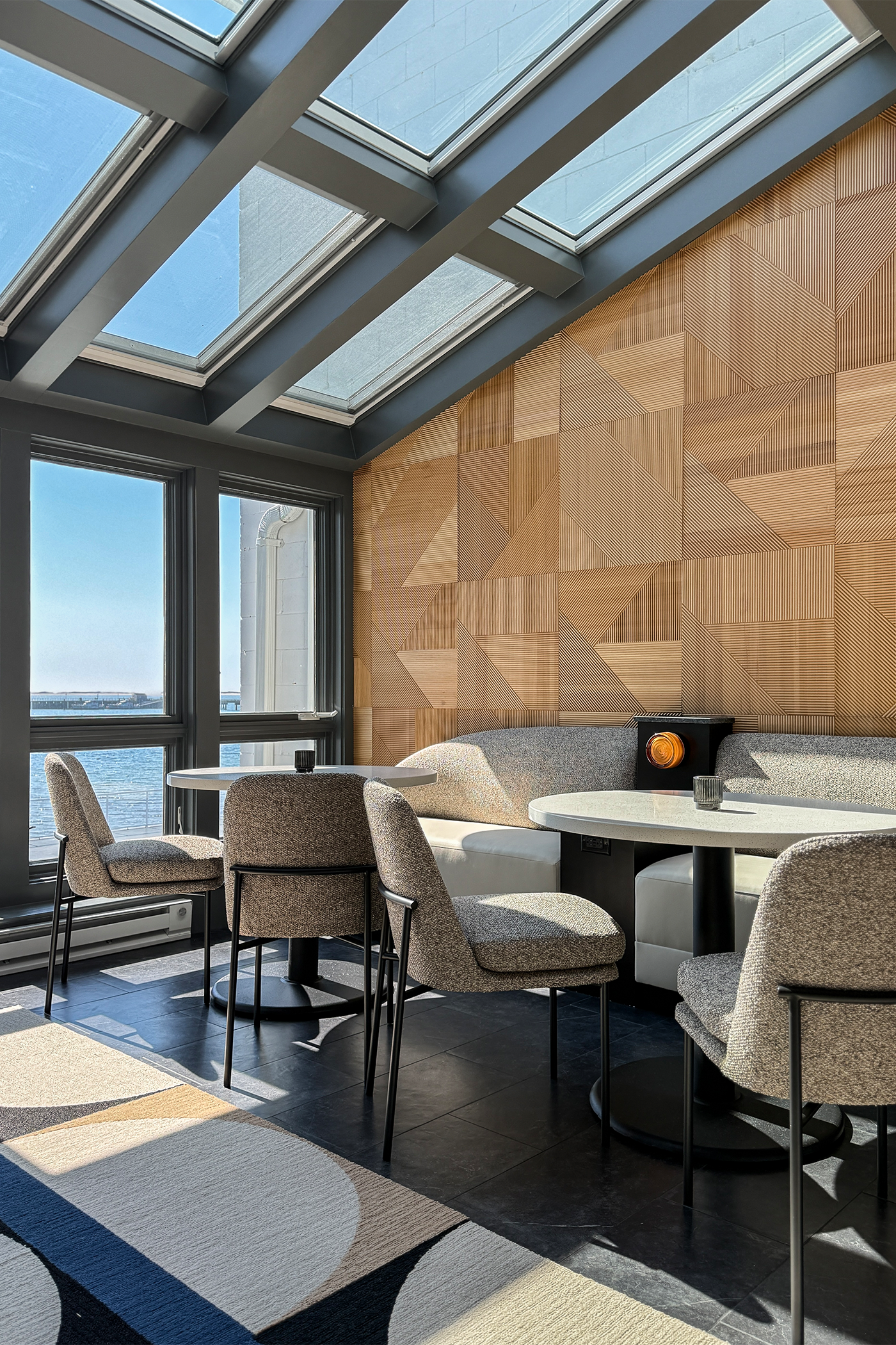 An interior space with large windows and a skylight, featuring a seating area with cushioned chairs, a round table, and a wall with a geometric wooden pattern.