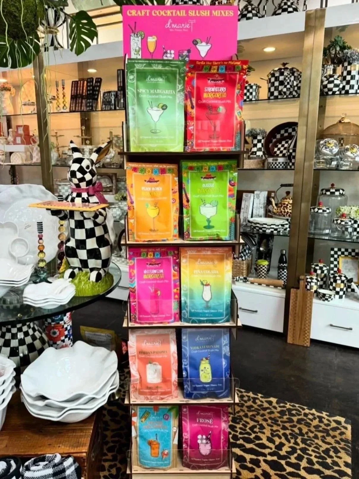 Display rack with colorful packets of craft cocktail slush mixes, including flavors like margarita, strawberry margarita, peach, and frose, in a store decorated with black and white checkered items and a leopard print rug.