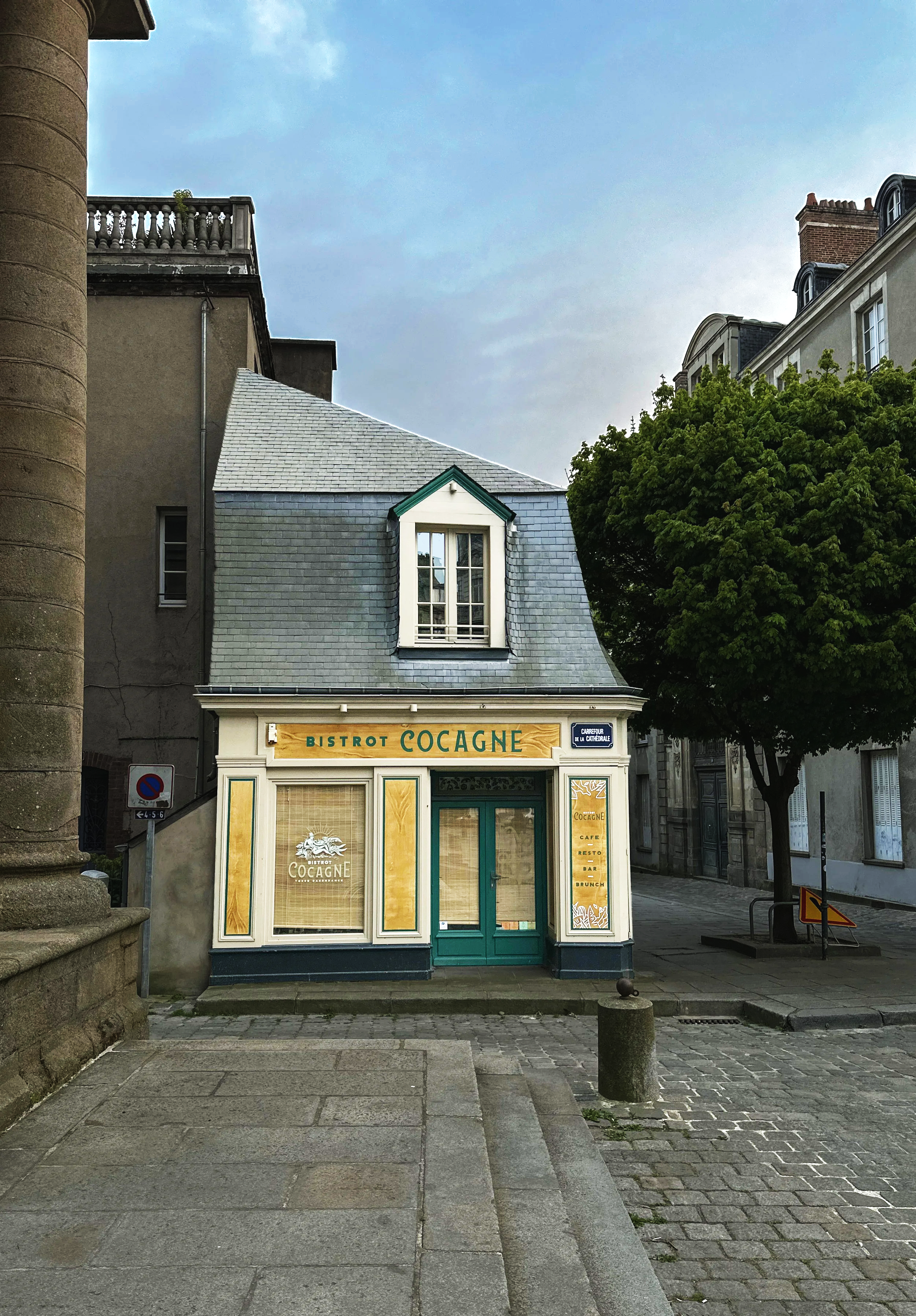 Small cafe or bistro named 'Cocagne' with beige storefront and green door, situated on a cobblestone street, with trees and older buildings behind it.