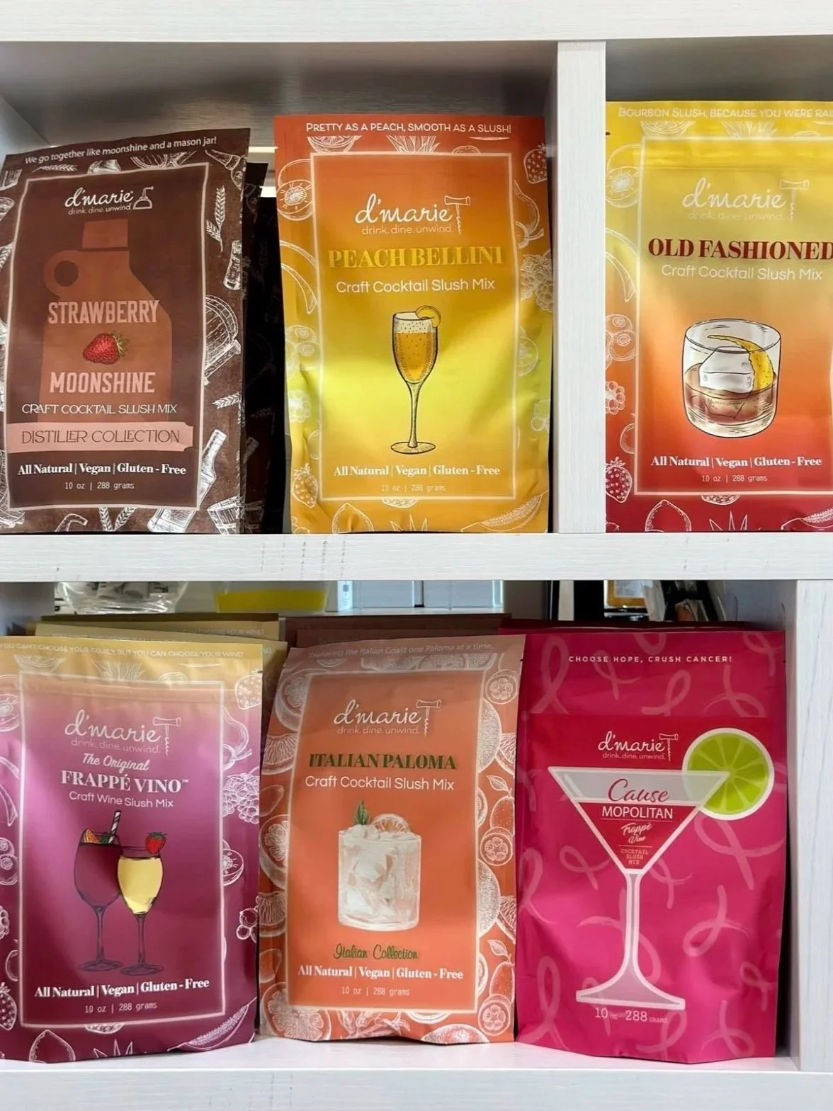 Display of colorful craft cocktail slush mix packets on a white shelf, featuring flavors like Peach Bellini, Old Fashioned, Frappé Vino, Italian Paloma, and Cause M'Opolitan, with illustrations of cocktails and text highlighting all-natural, vegan, and gluten-free ingredients.