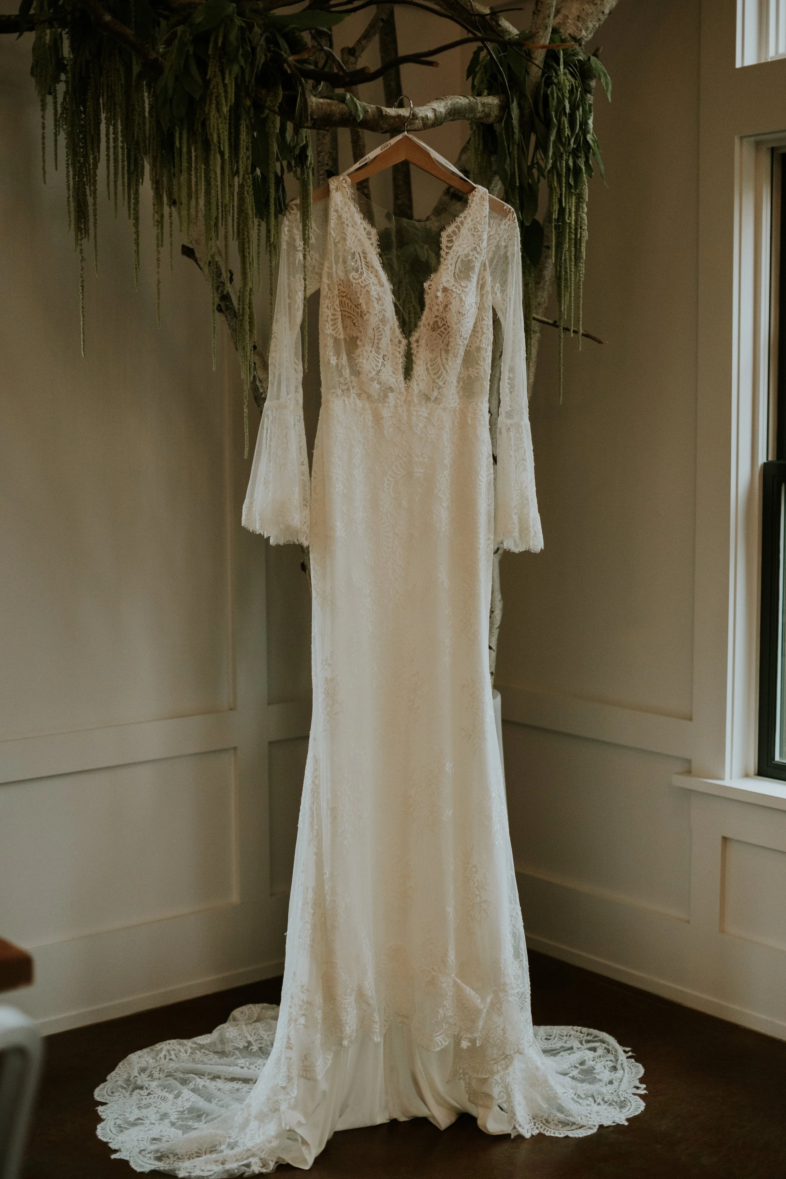 A white lace wedding dress hanging on a wooden hanger from a tree branch in a room with wainscoting on the walls and a window to the side.