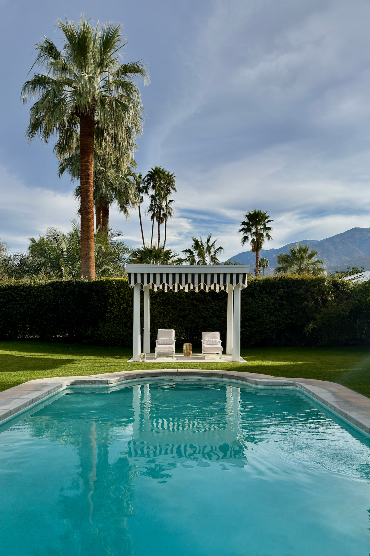 A backyard with a swimming pool, two white lounge chairs under a white pergola with black-and-white striped fabric, surrounded by green grass and tall palm trees, with mountains in the distance and a partly cloudy sky.