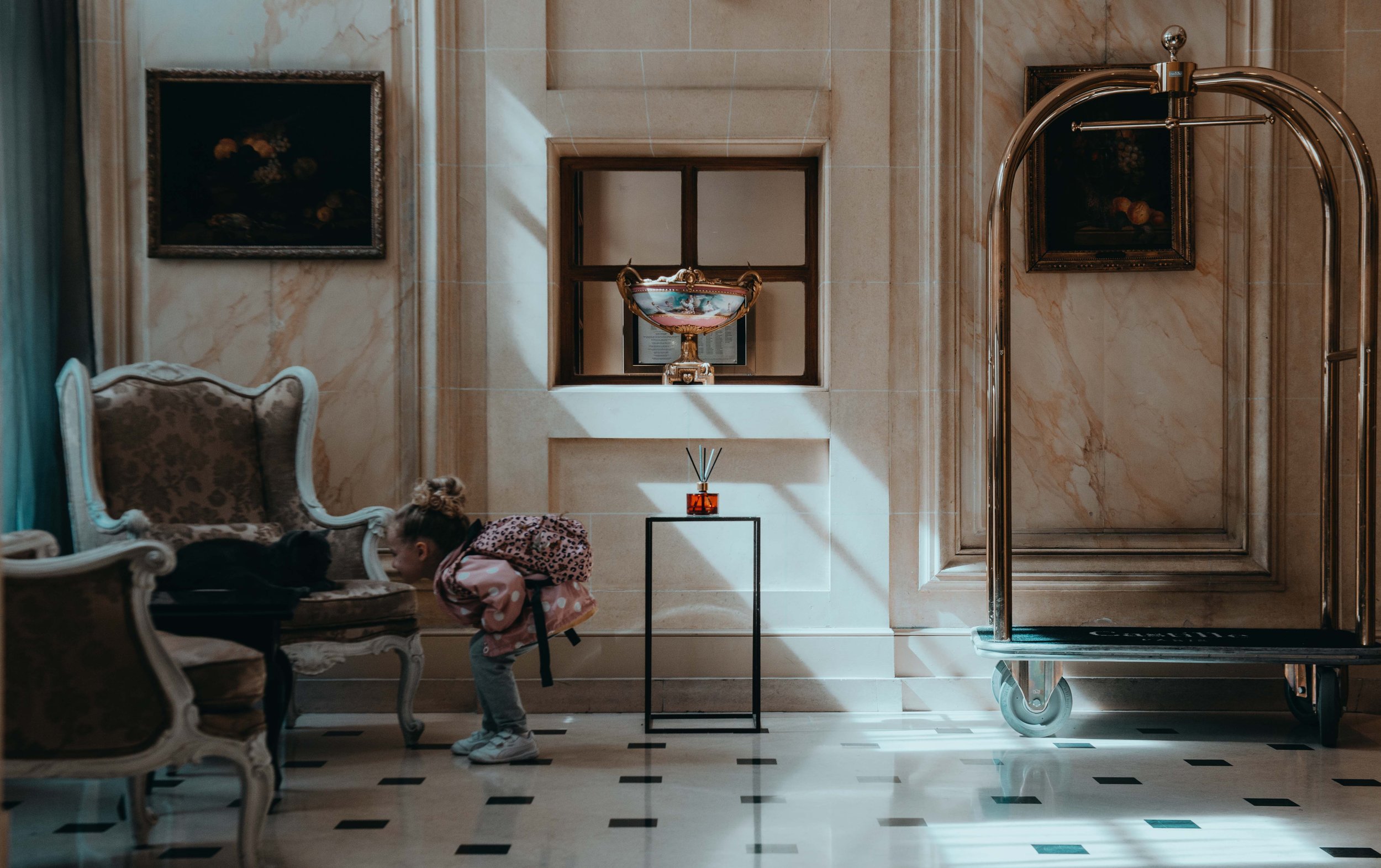 A young girl leans towards a black cat on a luxurious sofa in a grand hotel lobby.
