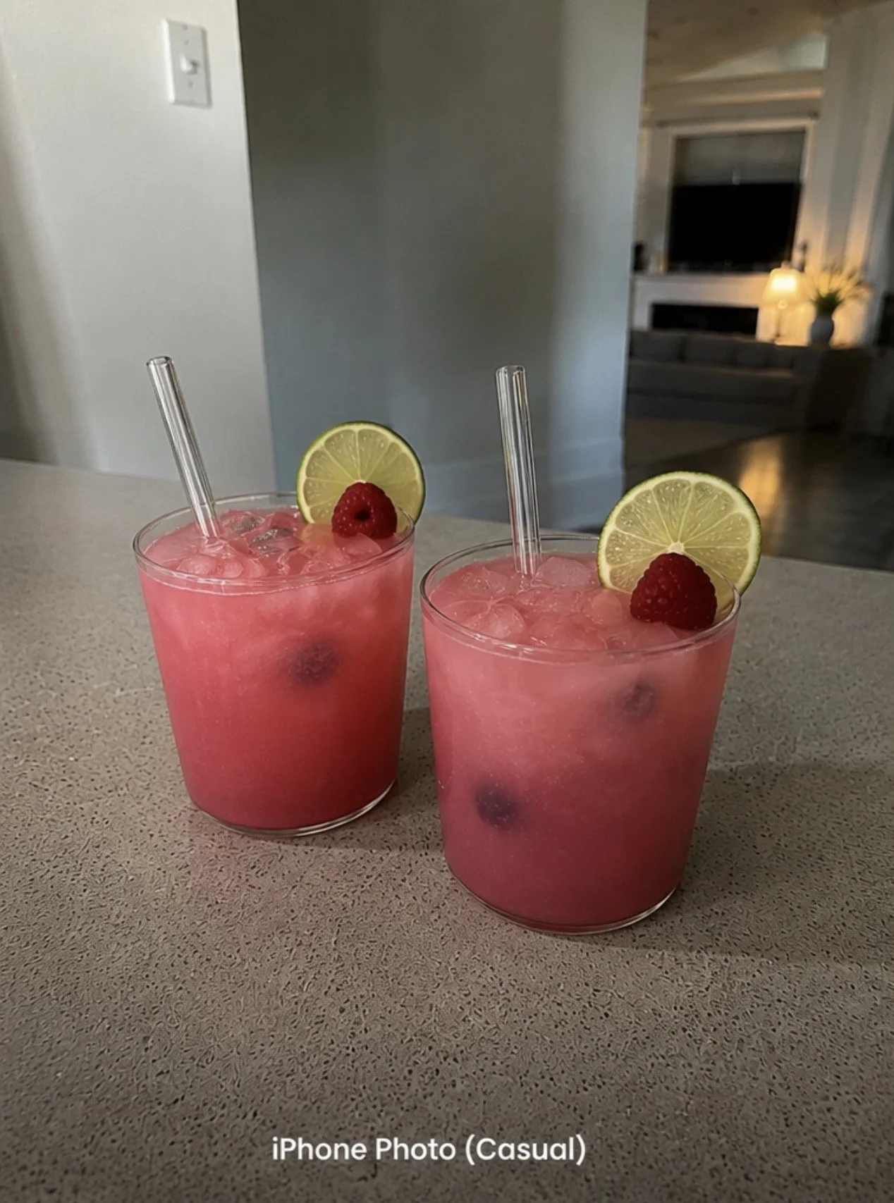 Two pink cocktails with lemon slices and raspberries on a beige countertop in a casual home setting.