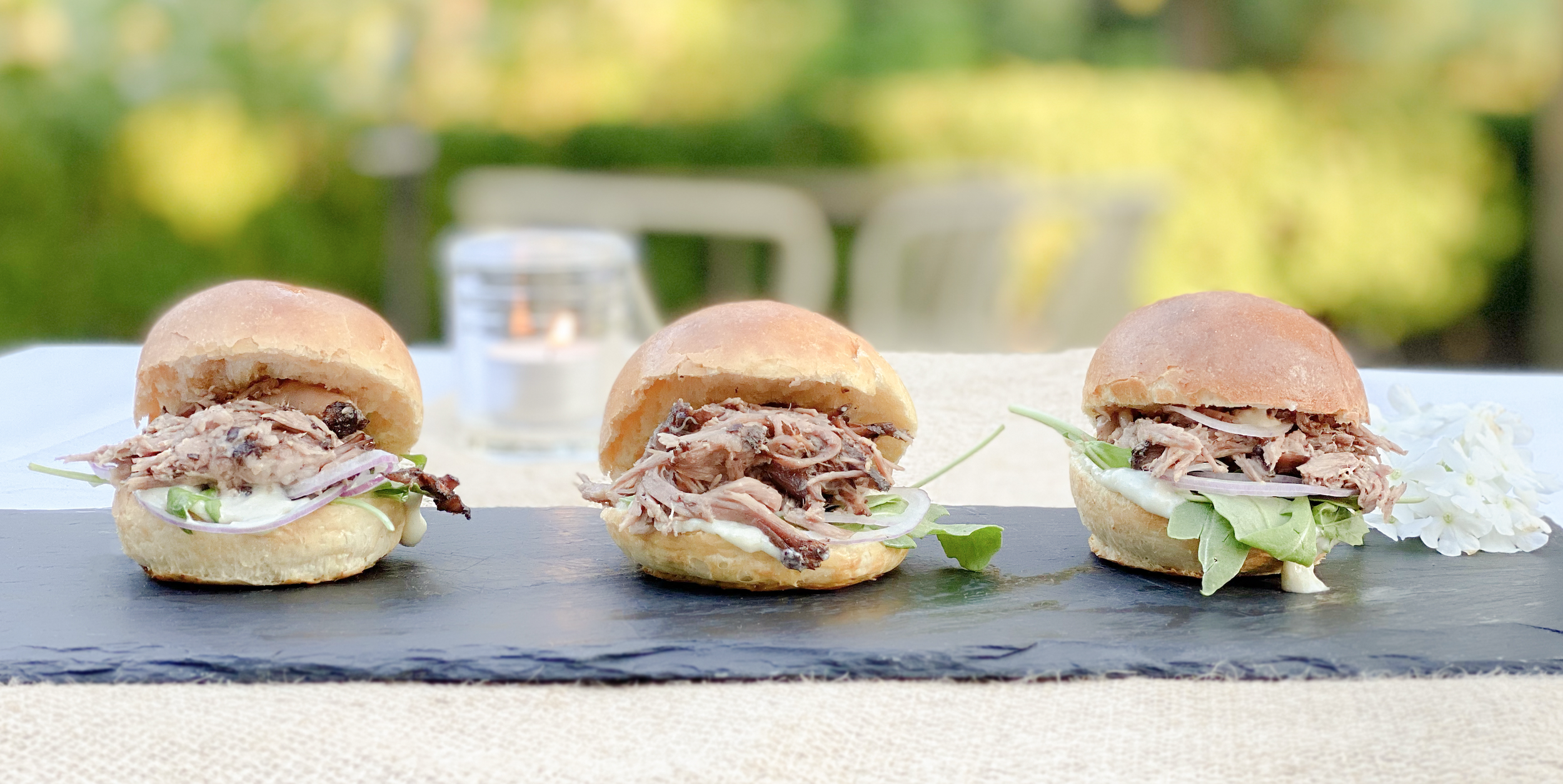 Three pulled pork sandwiches on a black slate serving board outdoors, with a blurred green background and some white flowers to the right.