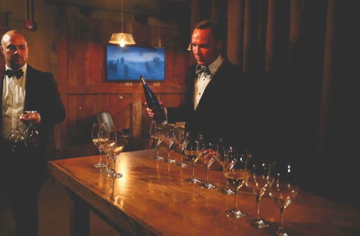 Two men in tuxedos preparing glasses of wine or champagne on a wooden table in a dimly lit room.