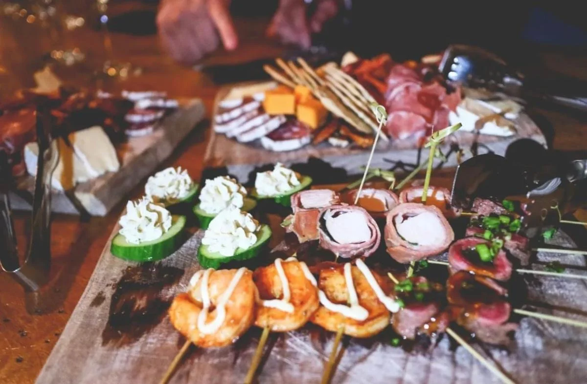 Assorted appetizers including skewered meats, cheese, and vegetables on a wooden platter, with slices of cured meats and cheeses in the background.