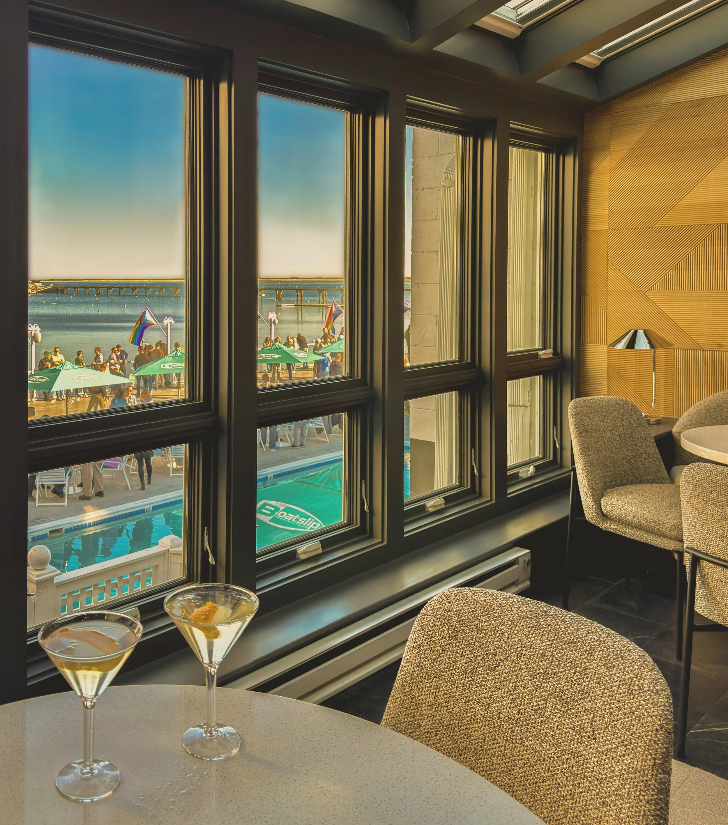Interior view from a room overlooking a beach scene with people, umbrellas, and rainbow flags through windows, with two martini glasses in the foreground.