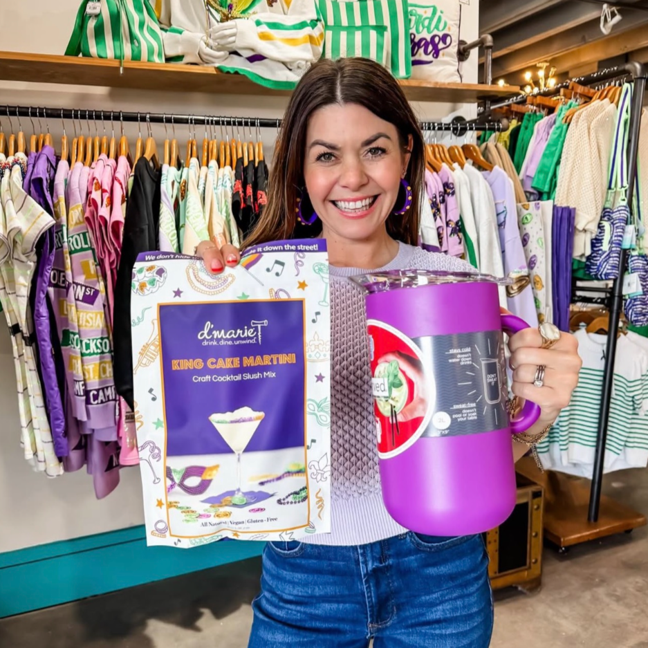 A woman smiling and holding a packet of King Cake Martini mix and a purple pitcher in a clothing store. Racks of colorful clothes, including shirts and sweatshirts, are visible in the background.