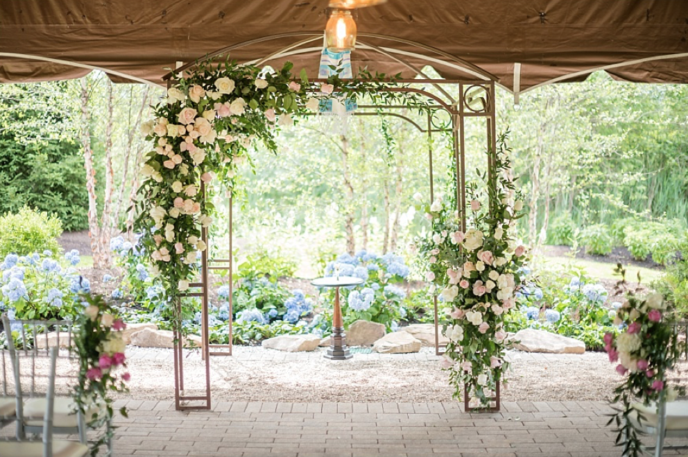 Outdoor wedding altar decorated with white and pink flowers, greenery, under a brown canopy, with blue hydrangeas and trees in the background.