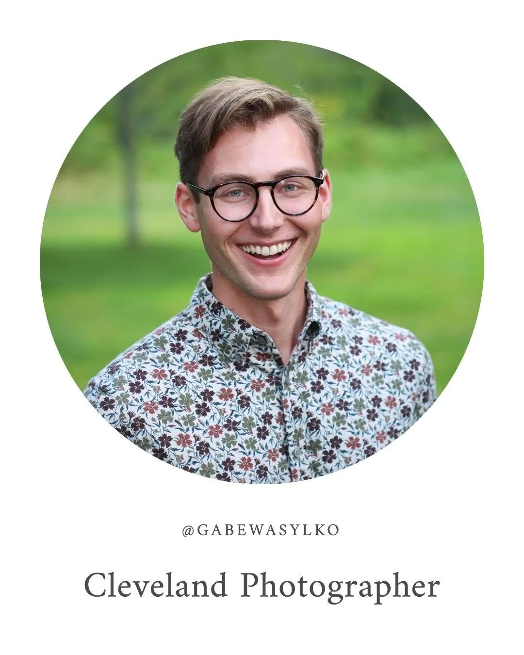 A young man with glasses, smiling, wearing a floral shirt, standing outdoors with a green background.