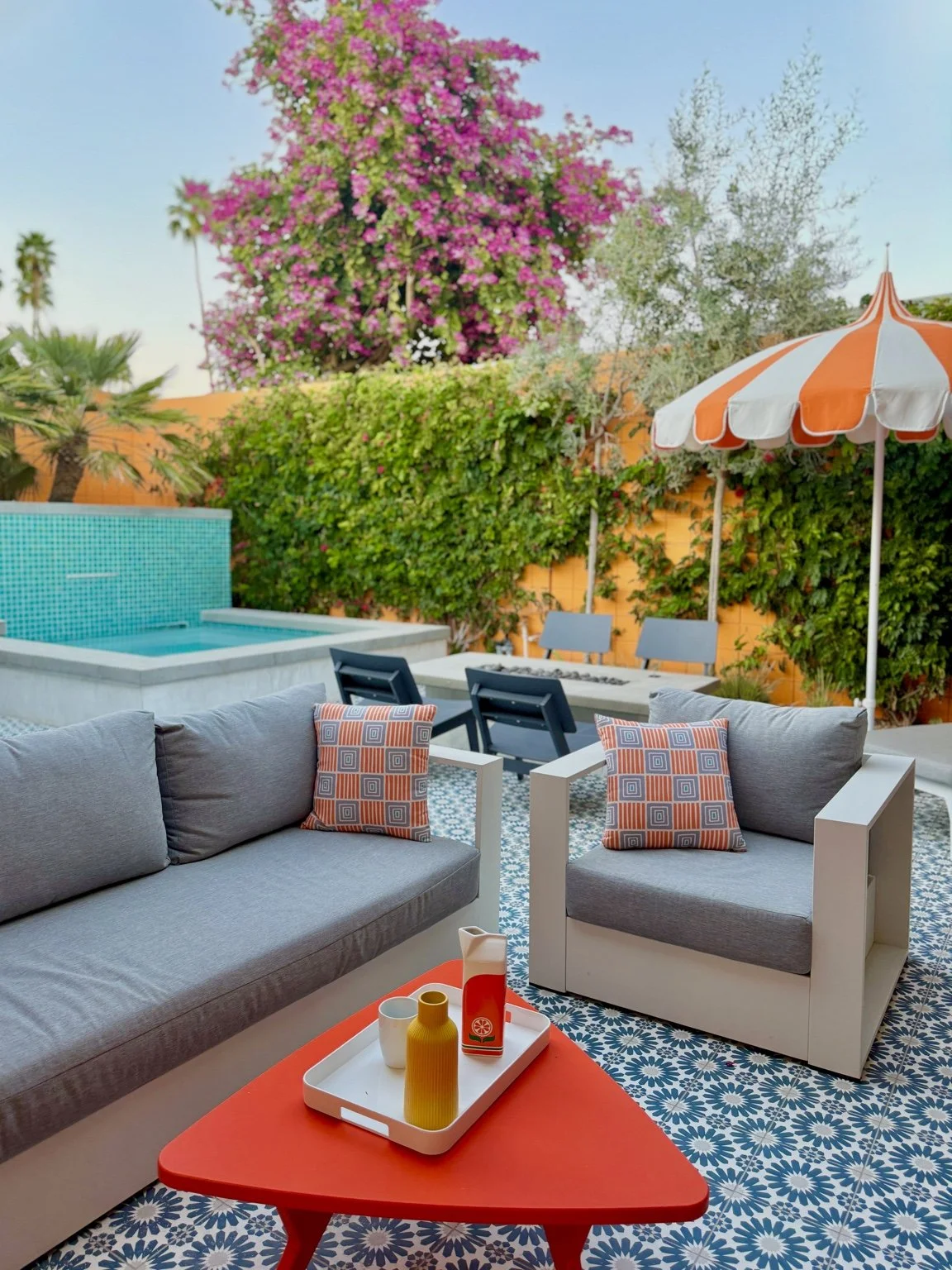 Outdoor patio with gray sofa and armchair, decorative pillows, red coffee table with vases and cups, patterned tile floor, umbrellas, a swimming pool, lush green plants, and pink flowering trees in the background.