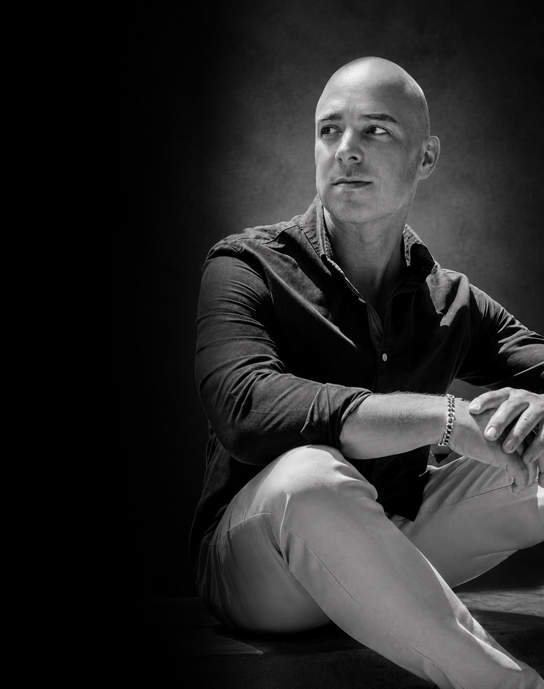 A black-and-white portrait of a bald man in a dark shirt sitting with one knee up, looking thoughtfully to the side.
