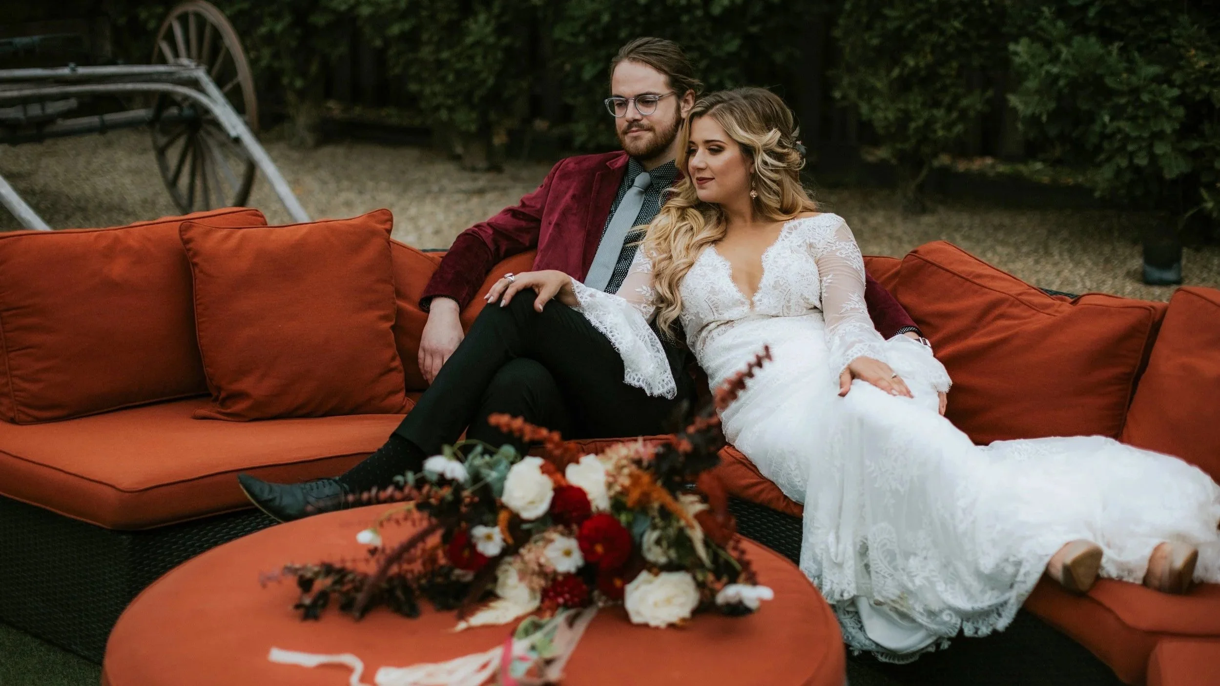 A man and woman sitting on an orange outdoor sofa, dressed in wedding attire, in a garden setting.