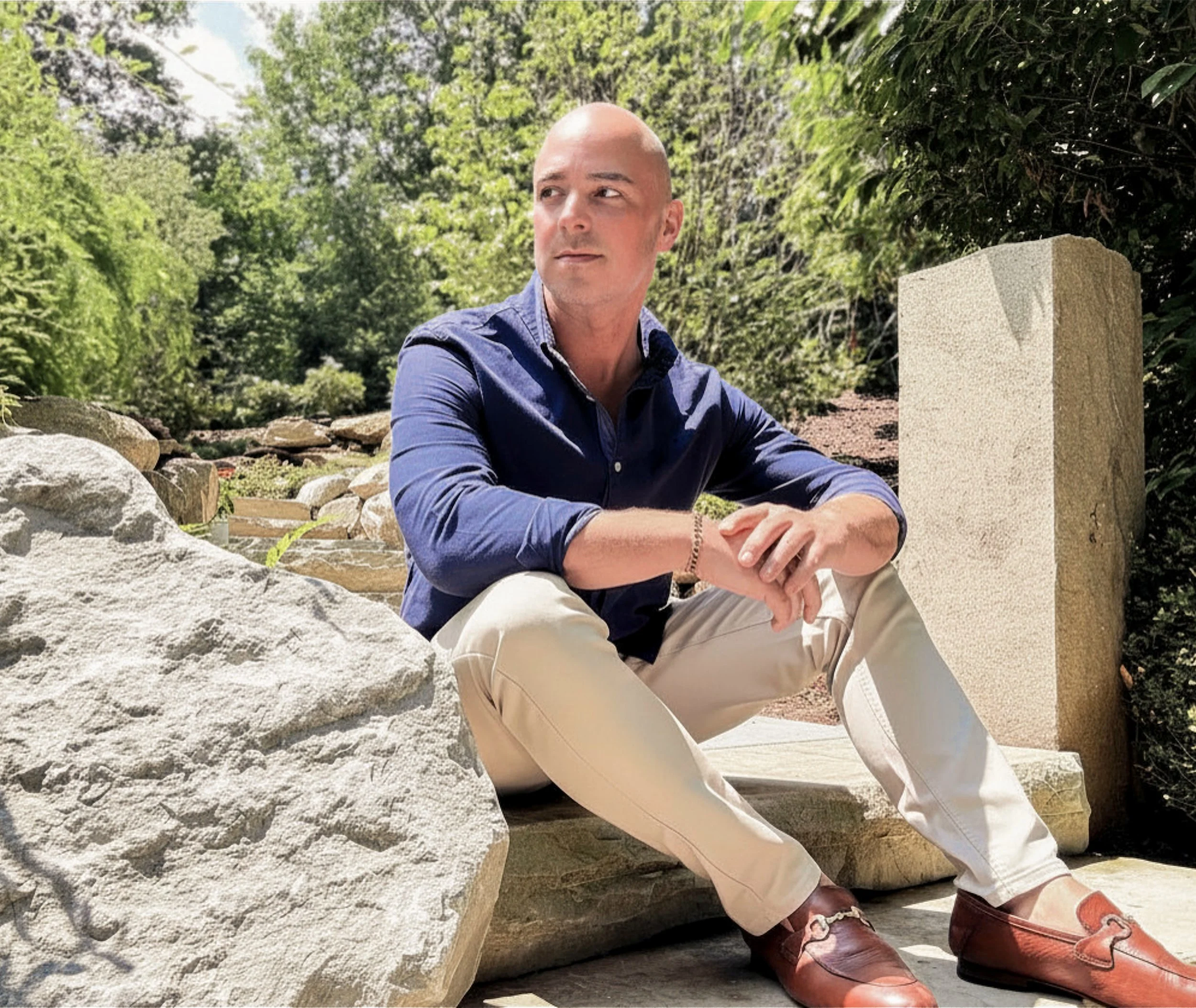A man with a shaved head and light skin sitting outdoors on stone steps, wearing a blue shirt, tan pants, and red shoes, with a background of green trees and bushes.