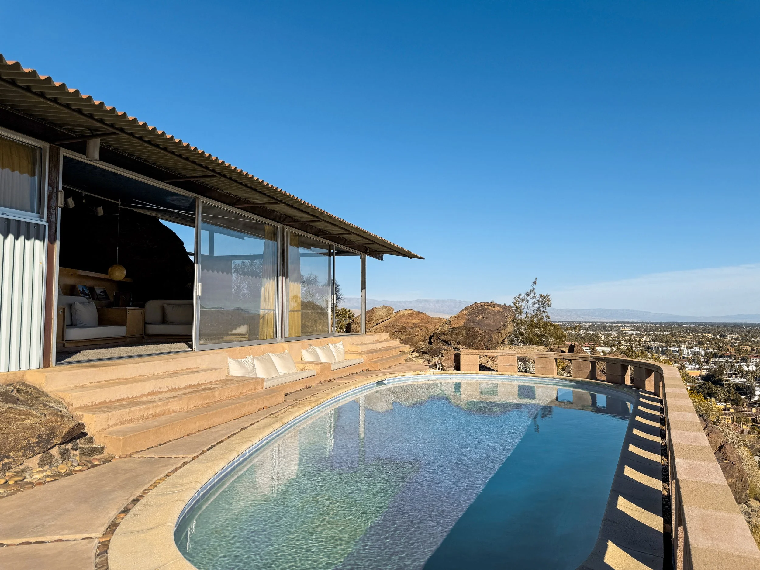 A house with large glass windows overlooking a swimming pool in a desert landscape with mountains in the distance.