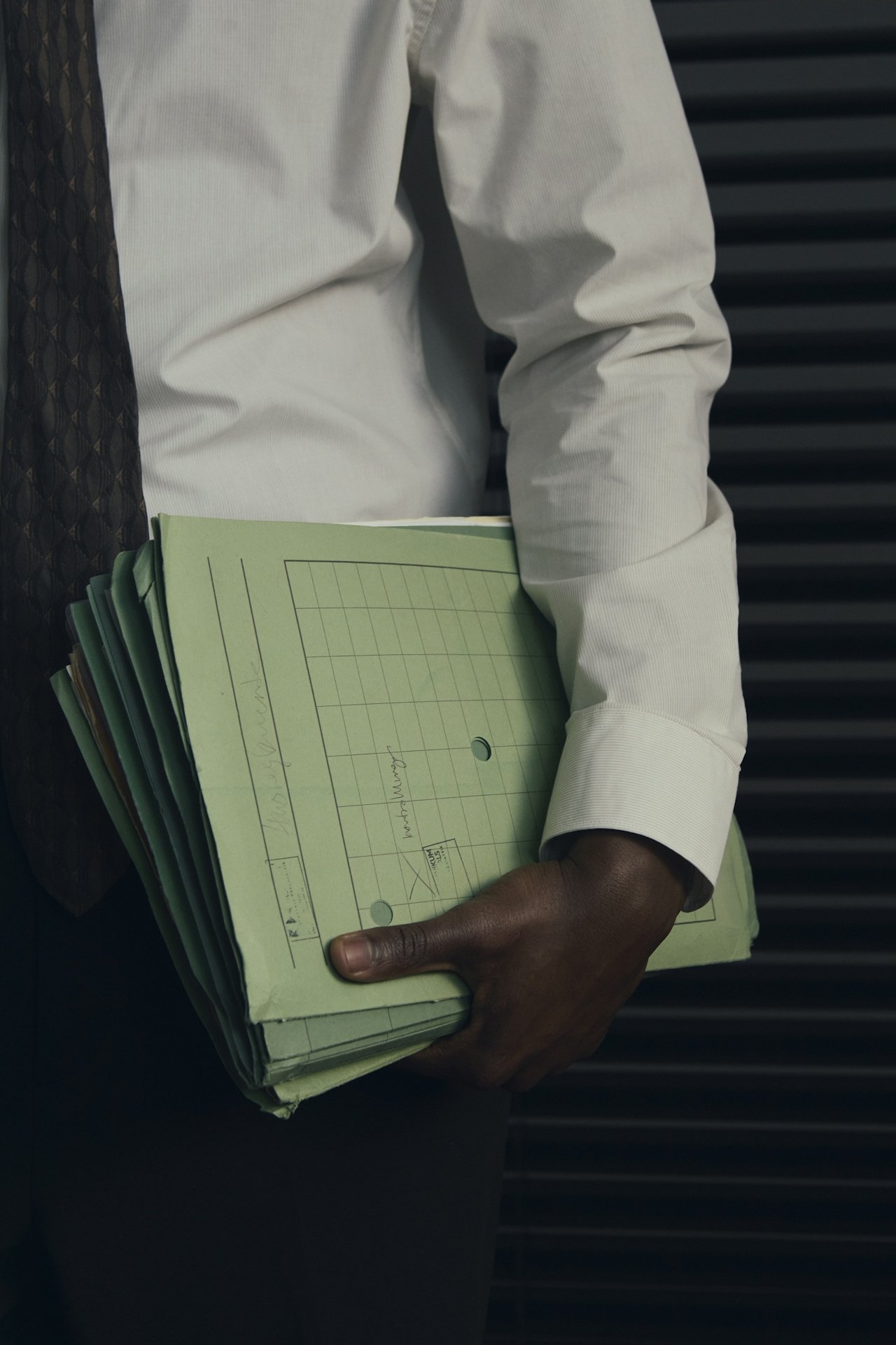 Person wearing a white shirt holding a stack of green notebooks or folders with handwritten notes, standing near a black, horizontal striped background.