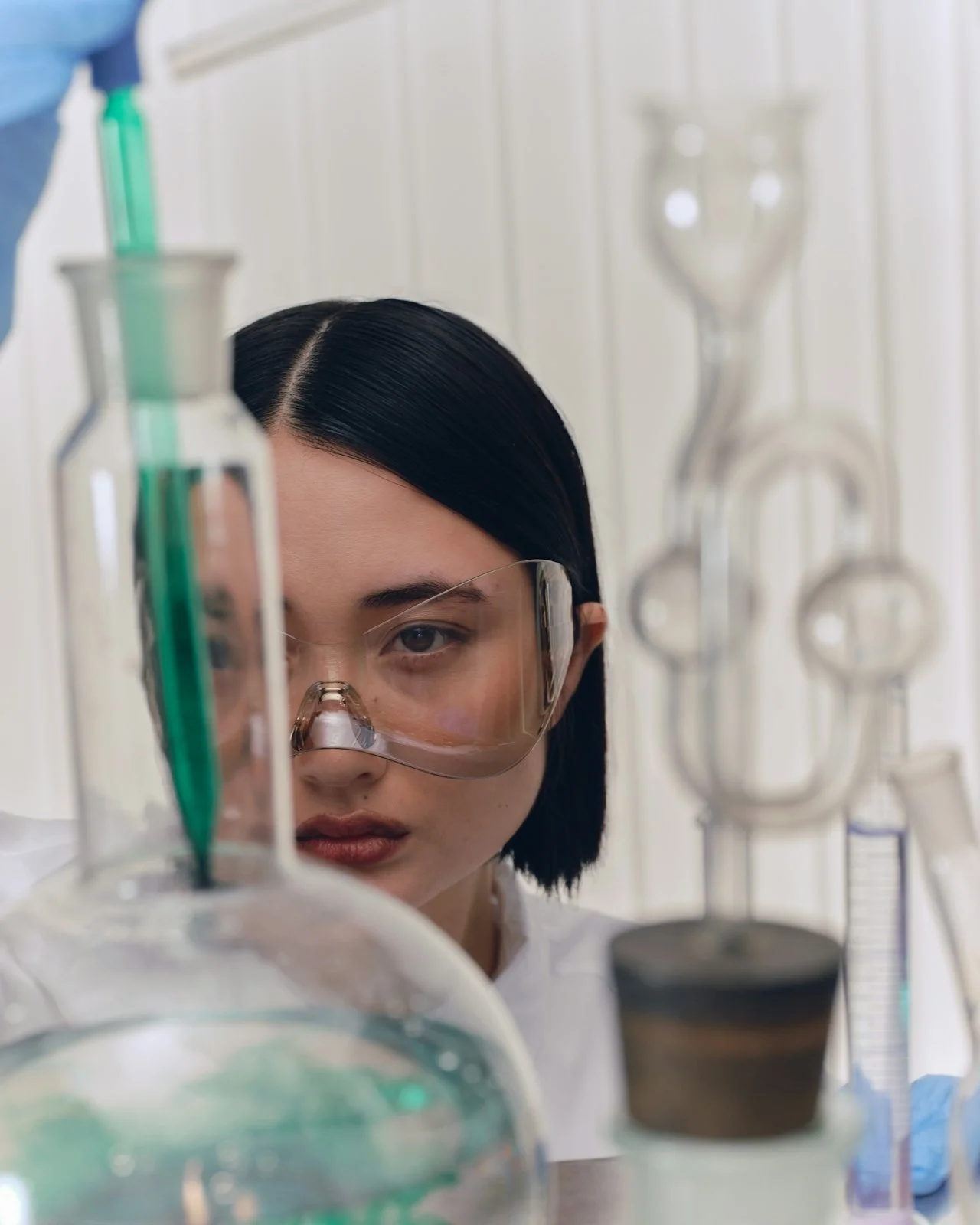A scientist with black hair and safety goggles looking at laboratory equipment including glassware and pipettes.