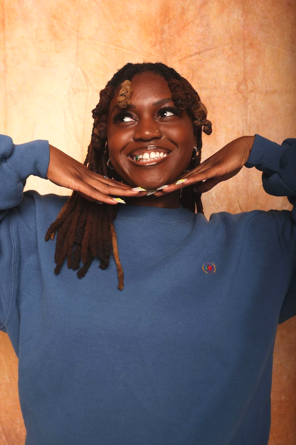 A woman with dark skin and long dreadlocks smiling and holding her hands under her chin, wearing a blue sweatshirt with a small logo, against a warm-toned background.