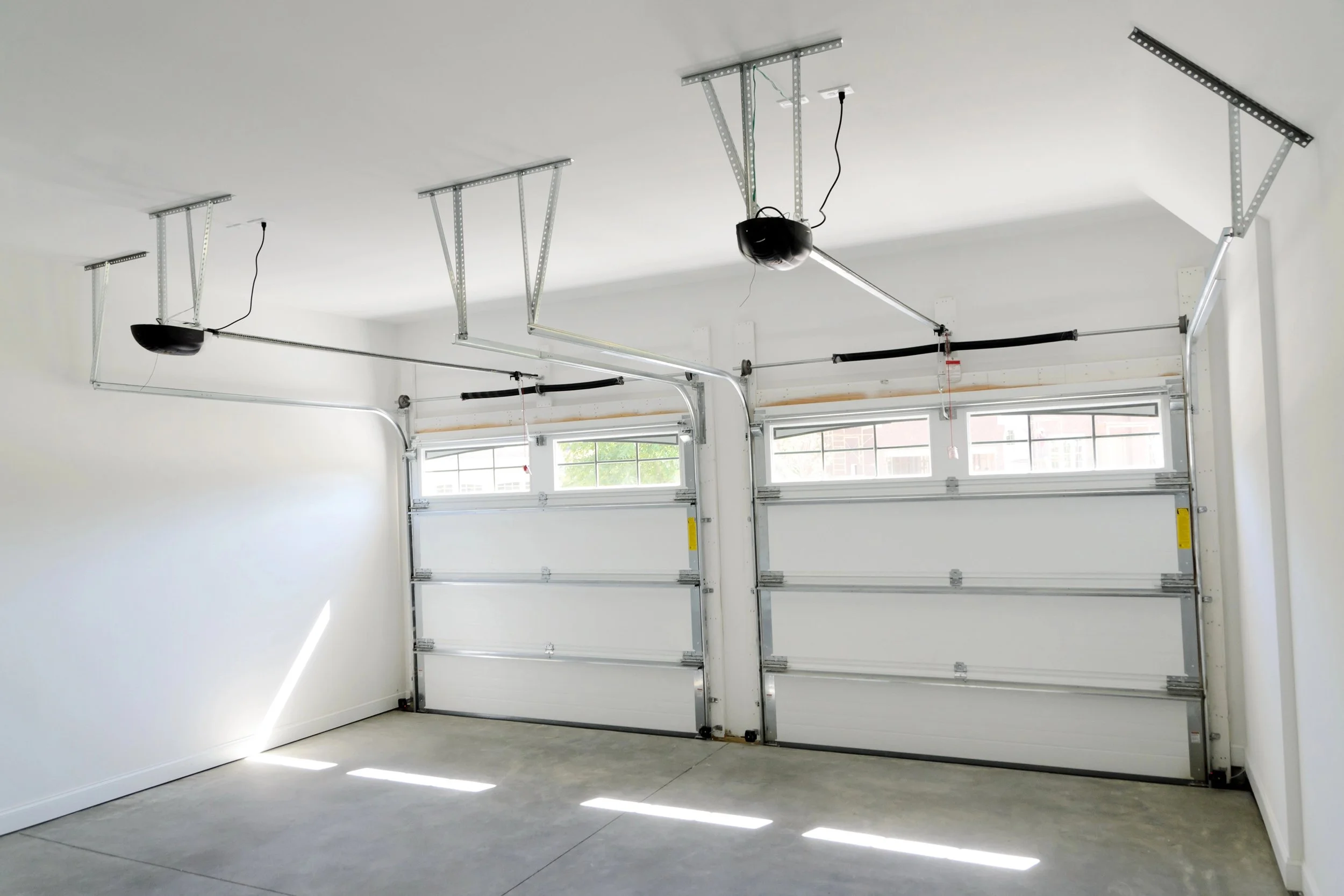 Empty garage with open sectional doors, garage door openers mounted on the ceiling, and sunlight streaming in through the windows.