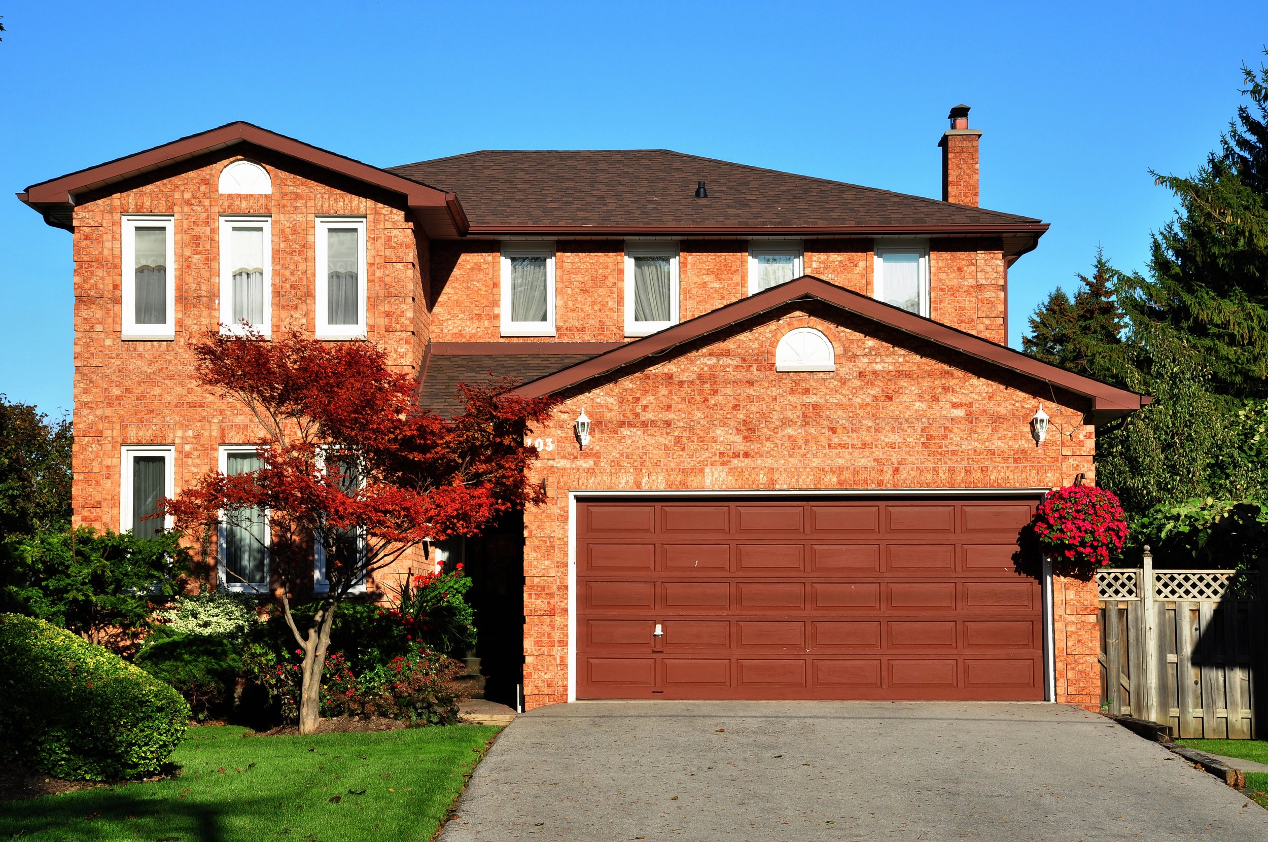 A red brick two-story house with a brown garage door, white window frames, a brick chimney, and a small front yard with green grass, trees, and bushes.