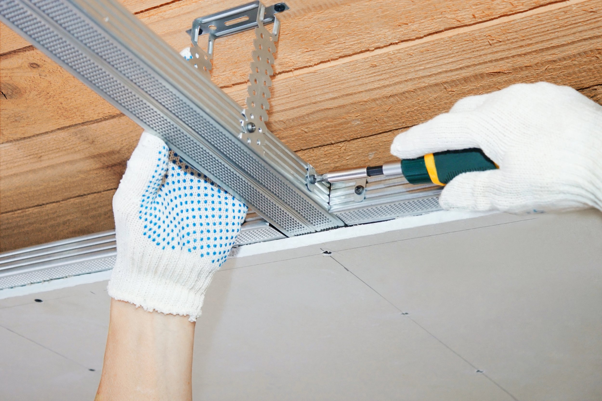 Person installing or working on a ceiling with drywall and metal framing, using a screwdriver while wearing white gloves.